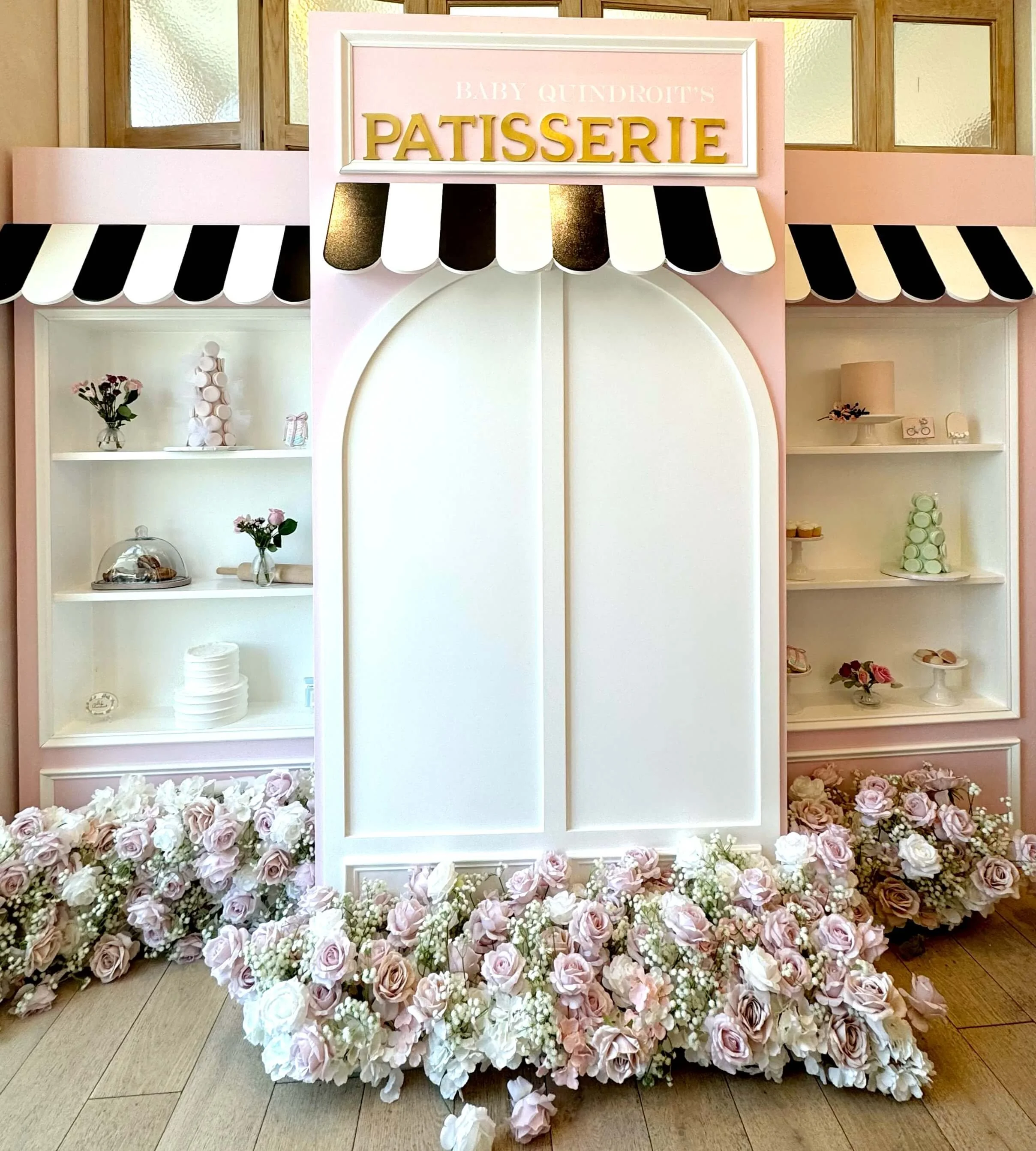Pink French patisserie dessert wall backdrop with floral arrangement, featuring “Baby Quindroit’s Patisserie” signage, pastel shelves, macarons, cakes, and a bakery-themed baby shower display.