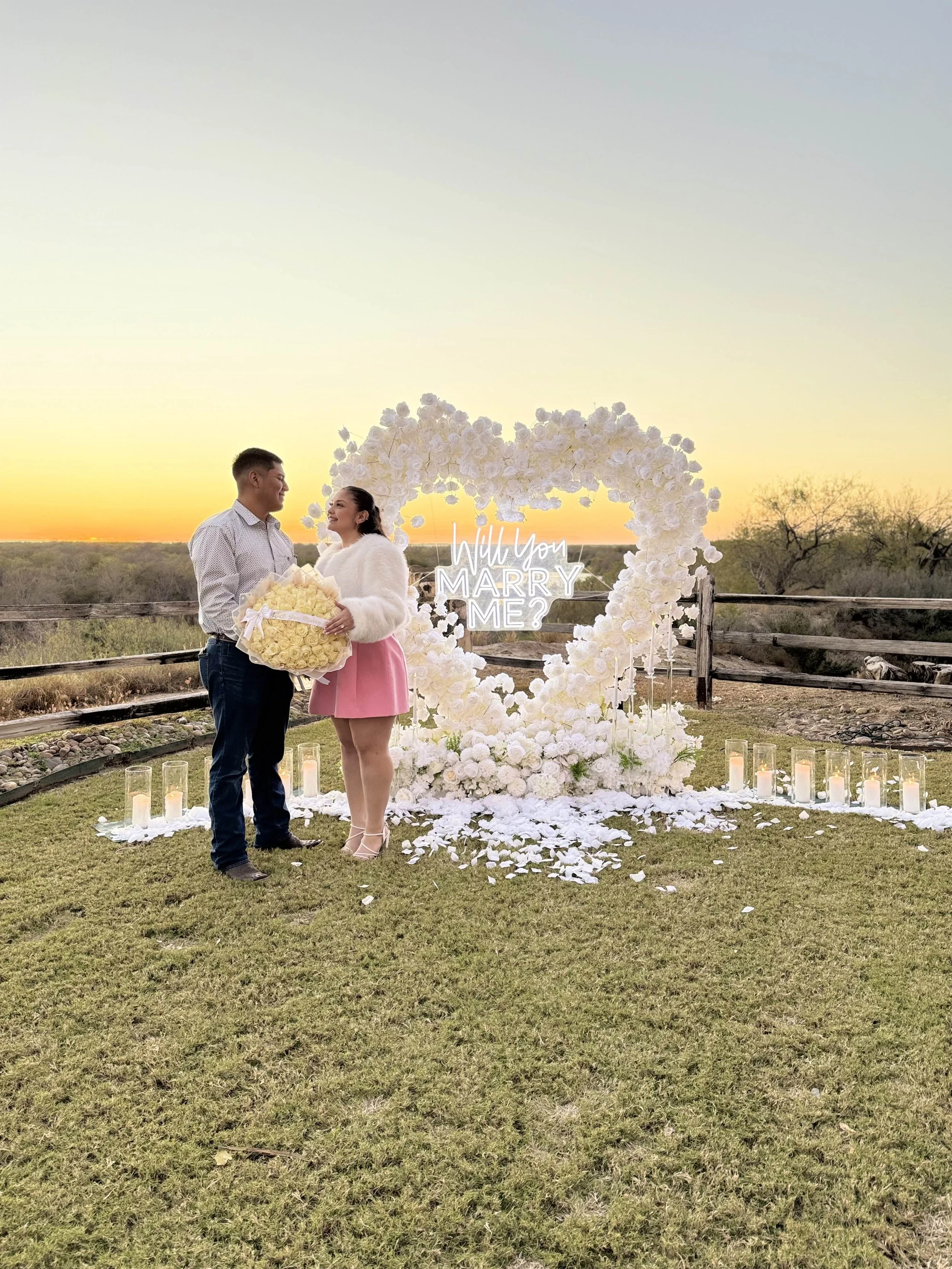 A couple standing outdoors at sunset, with a large heart-shaped floral decoration behind them, and a neon sign that says 'Will you marry me?' The woman is holding a large bouquet of yellow roses, and the man is smiling at her. Candles are placed on t