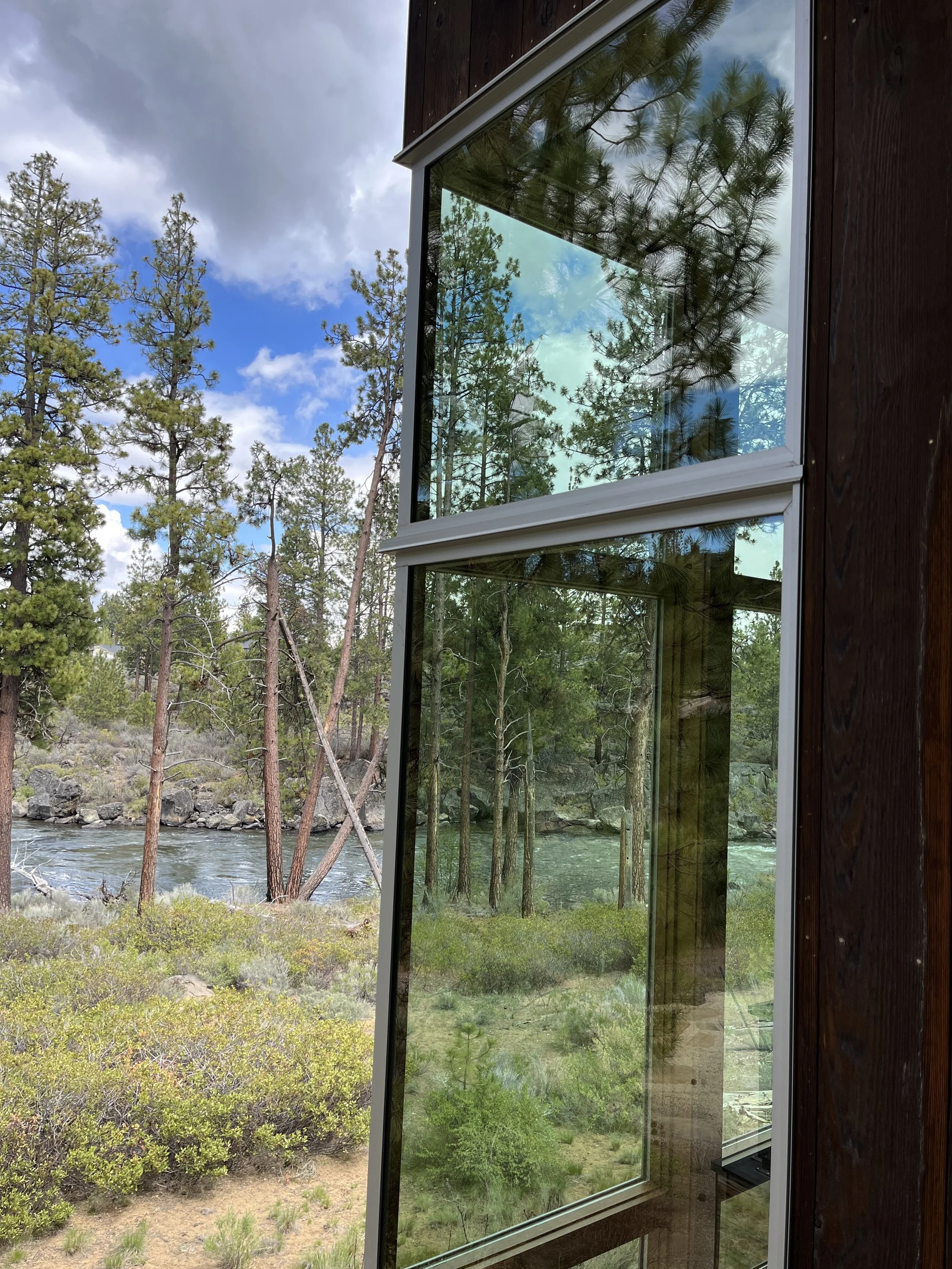 Reflection of trees and sky in a large window of a wooden house located in a forested area near a river.