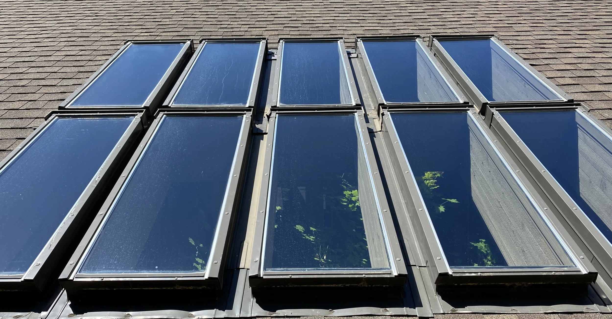 Photograph of multiple skylights installed on a sloped rooftop, with visible metal frames and glass panels reflecting the sky.