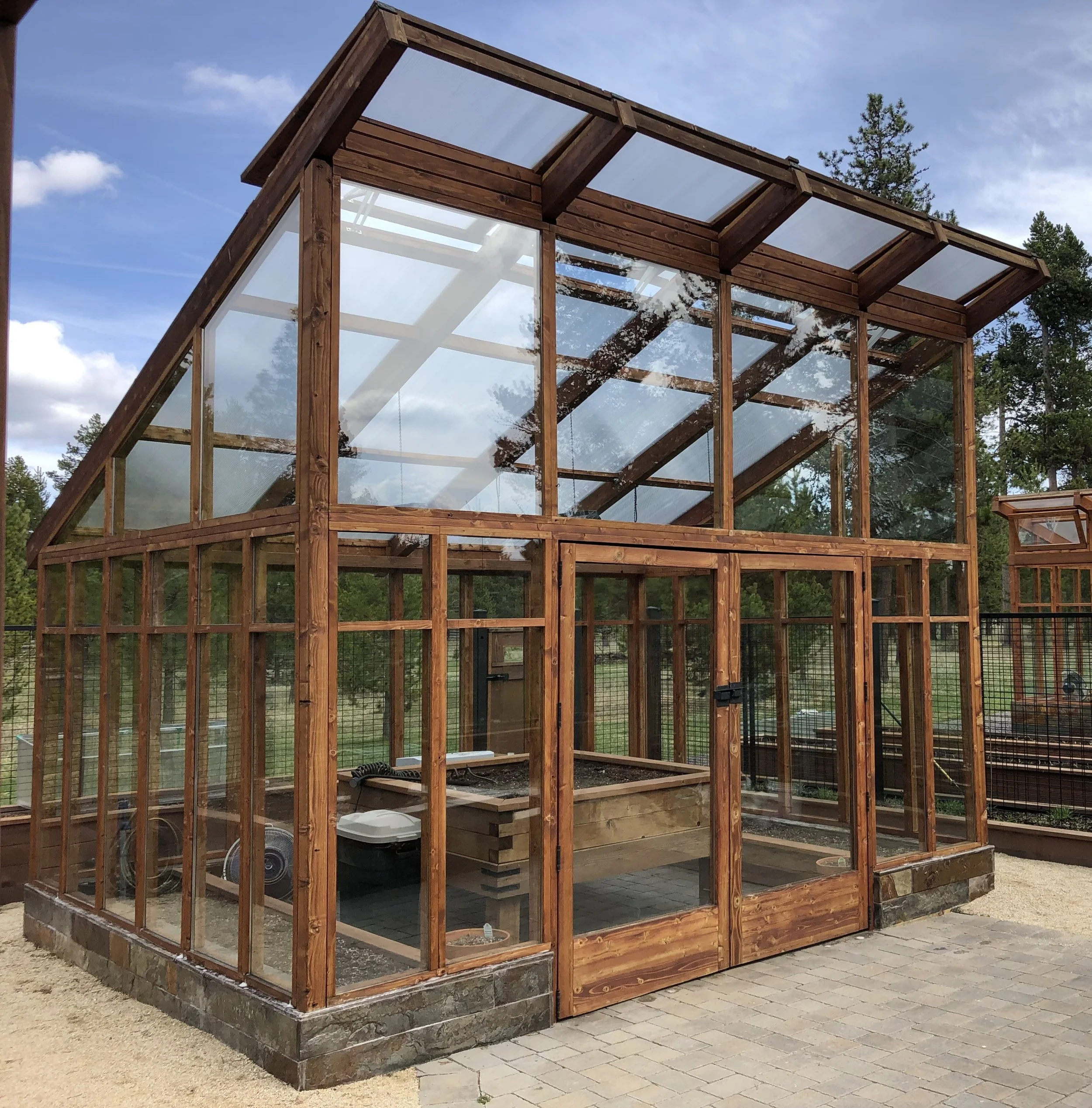 A wooden greenhouse with glass panels, situated on a paved area outdoors, surrounded by trees and a wooden fence.