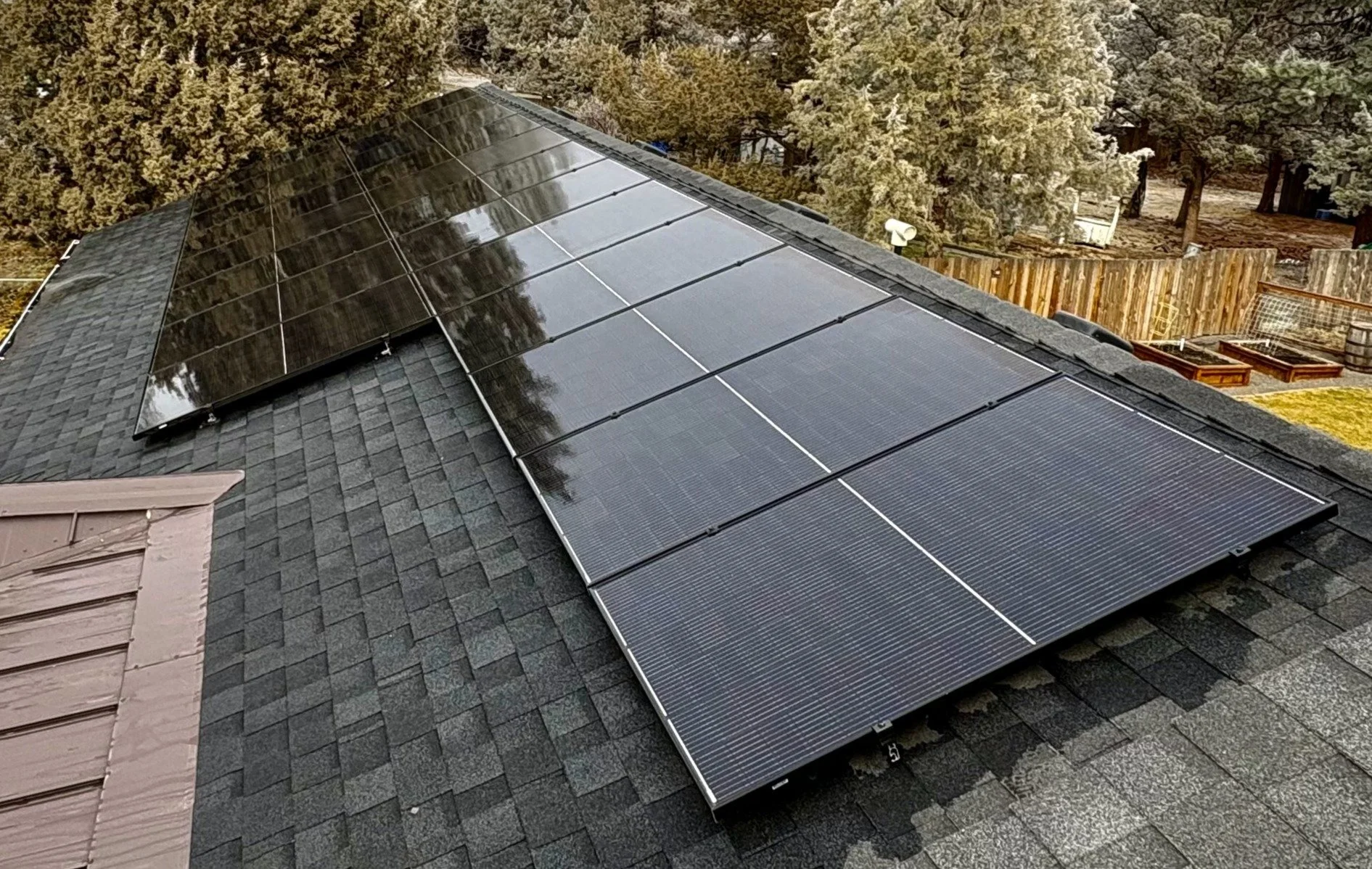A rooftop with multiple black solar panels installed on dark gray shingles, with trees and a wooden fence in the background.