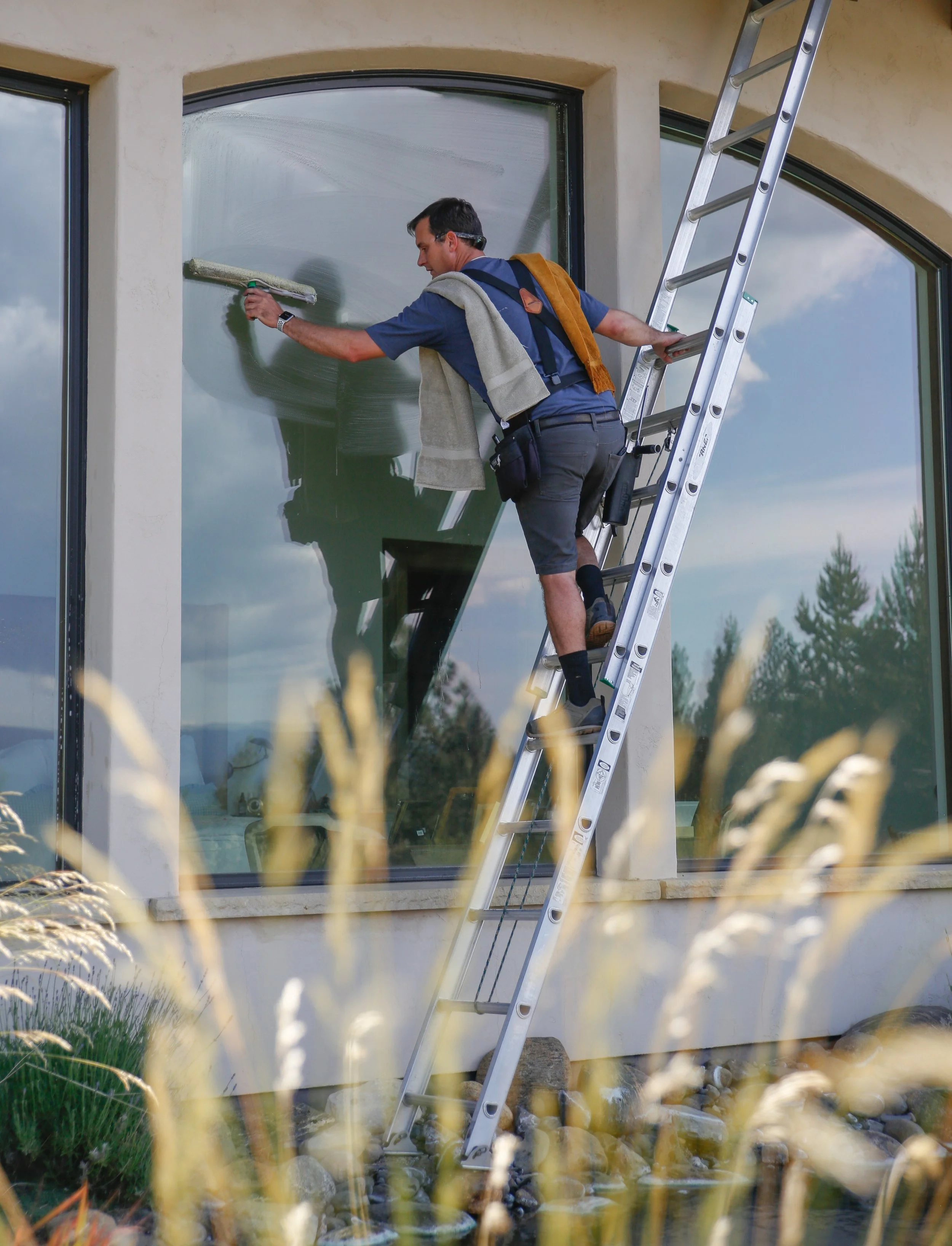 A man cleaning a large window of a building with a roller brush, standing on a ladder.