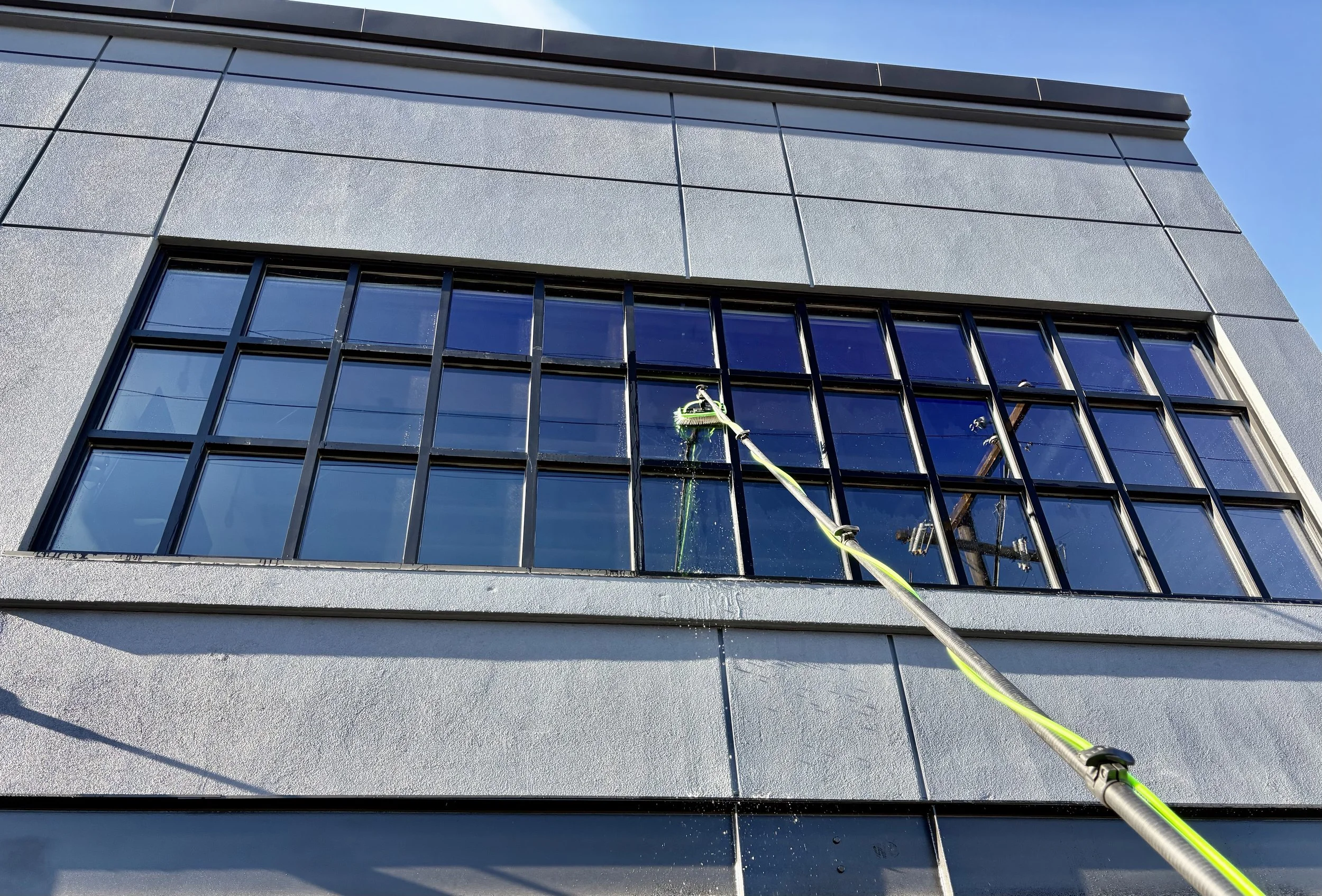 Worker cleaning exterior window on a modern building using a long pole and brush.