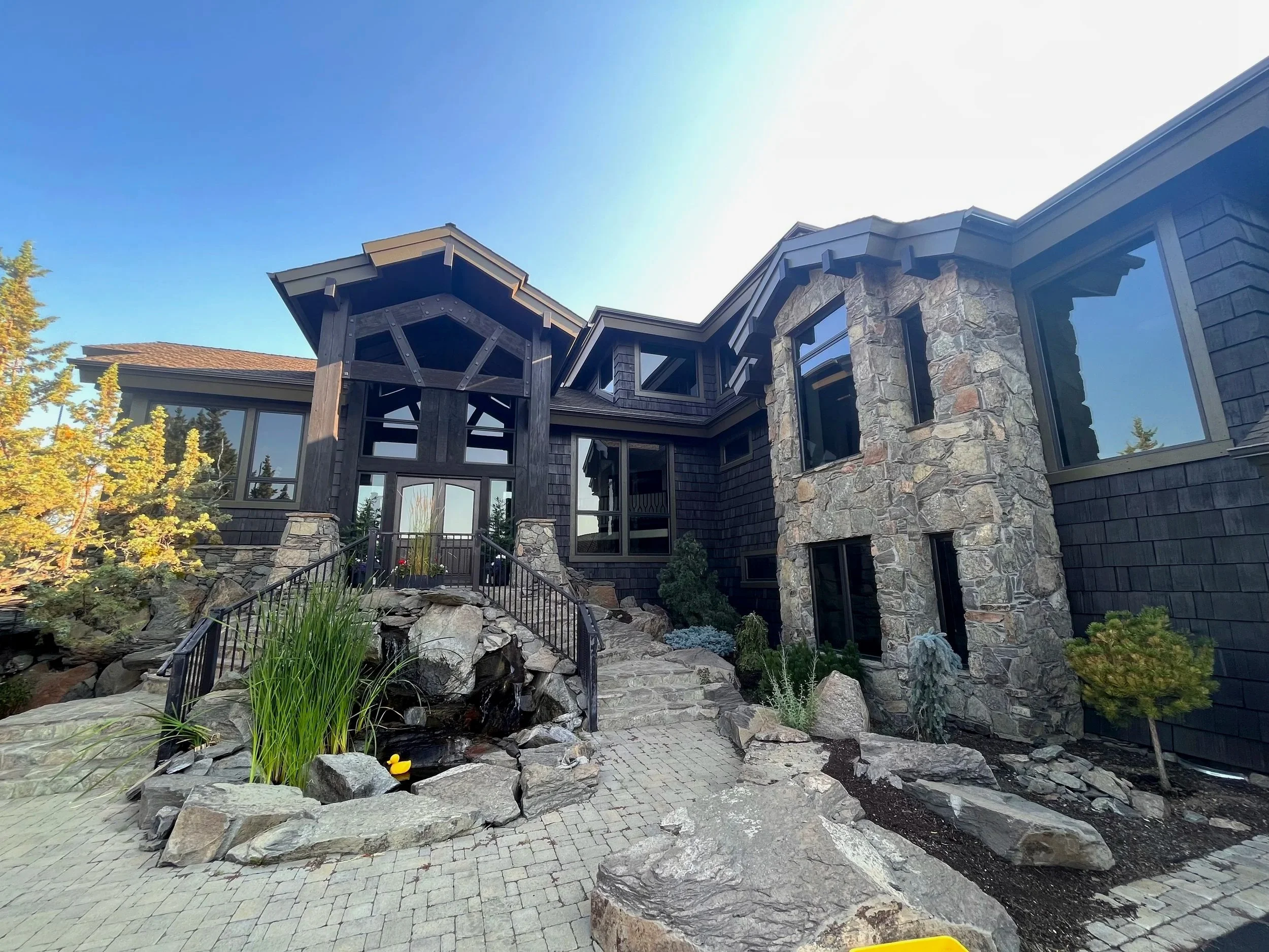 A modern house with a stone and dark wood exterior, large windows, a front porch with stairs, landscaped rocks, plants, and a small pond with a rubber duck, under a blue sky.