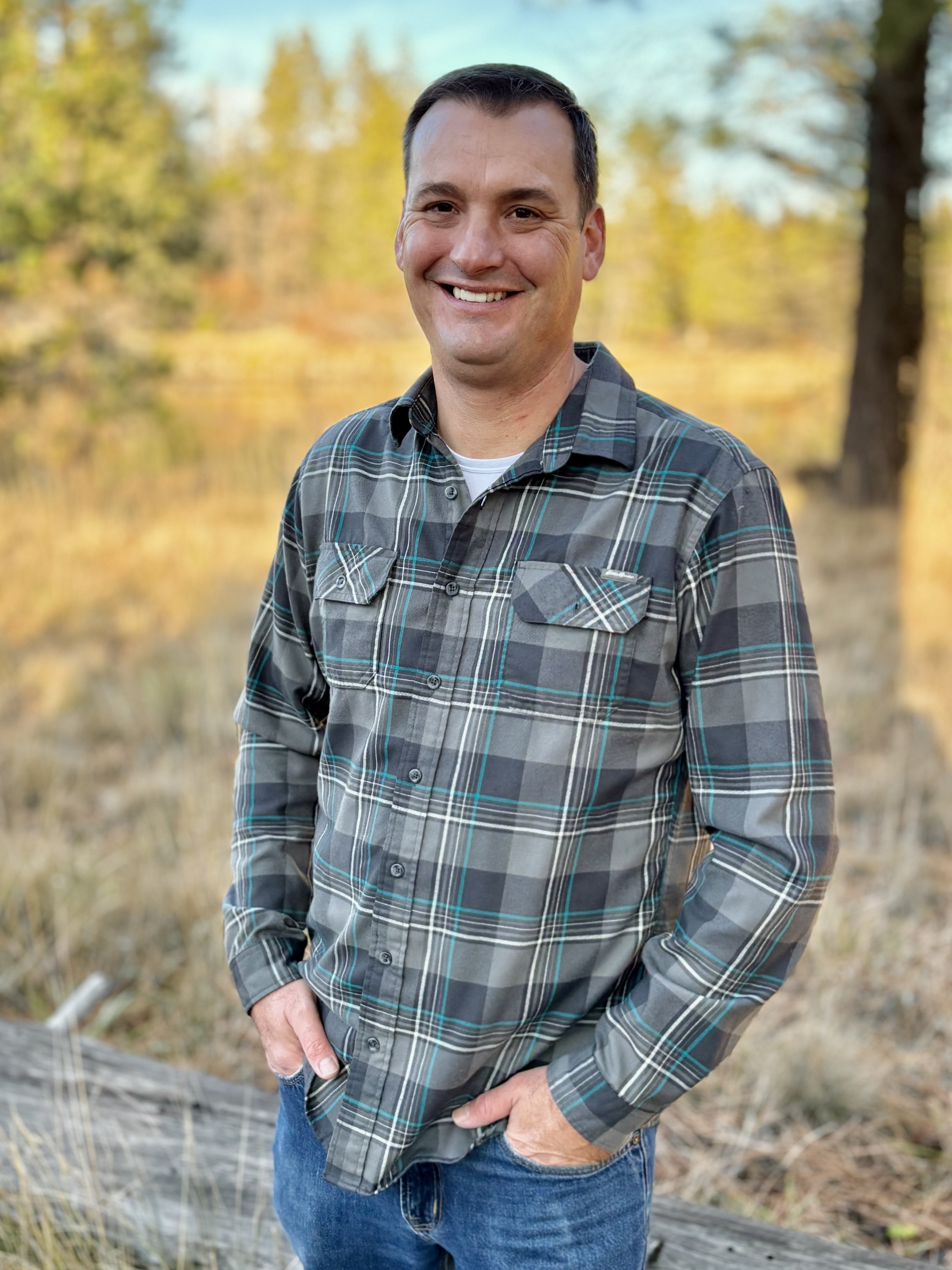 A man standing outdoors in front of a lake and trees with fall foliage, wearing a plaid shirt and smiling.