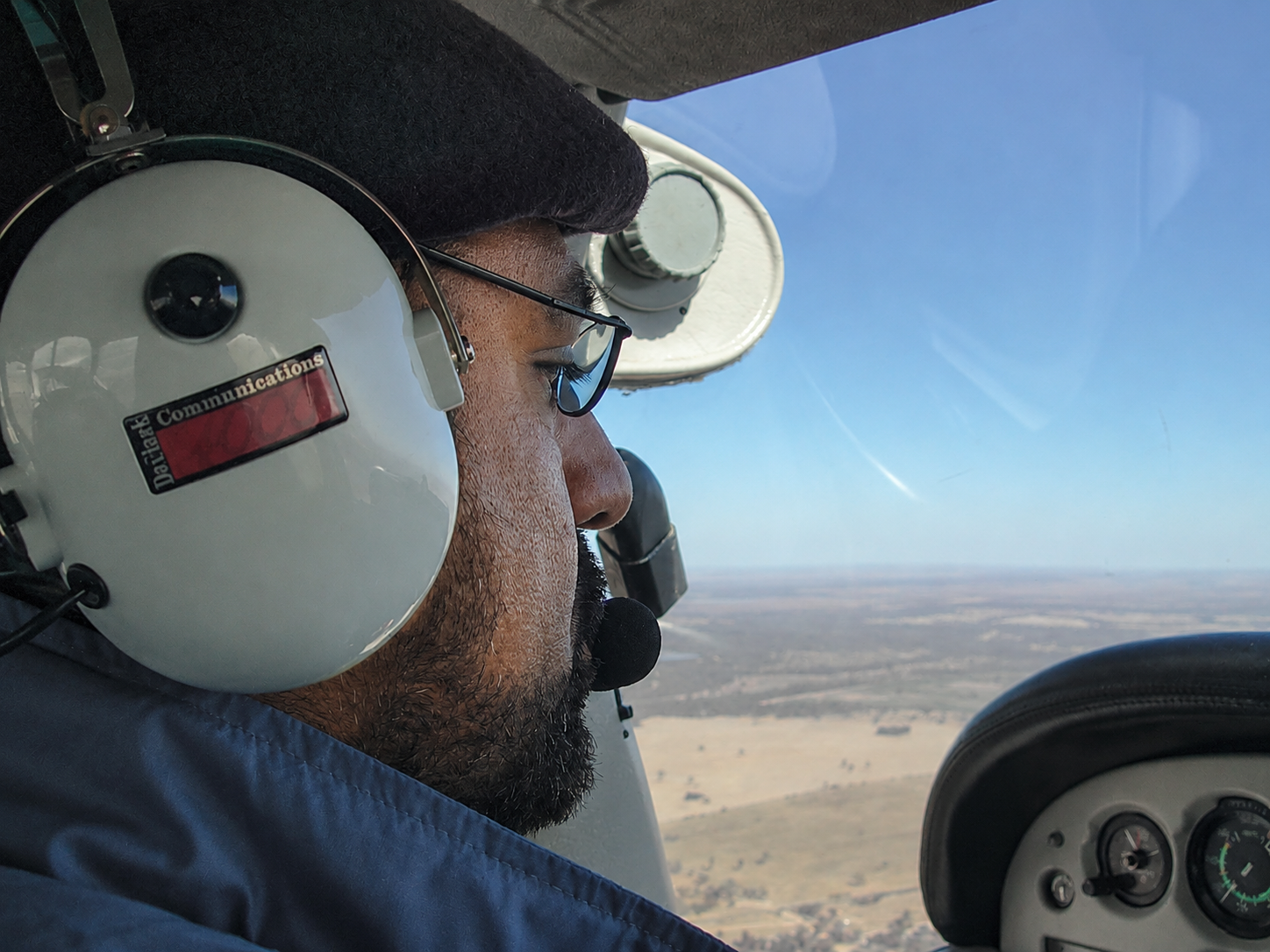 Close-up of Navnit Bhalla pilot wearing a white headset and sunglasses, flying a small airplane over a flat landscape.