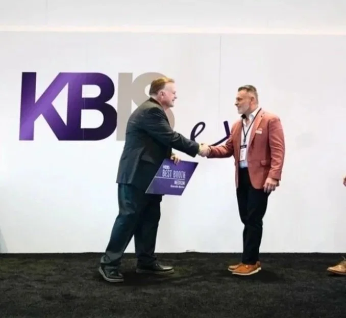 Two men shaking hands at a KBS event, with one holding an award plaque that says 'Best Booth.' The backdrop has the KBS logo.