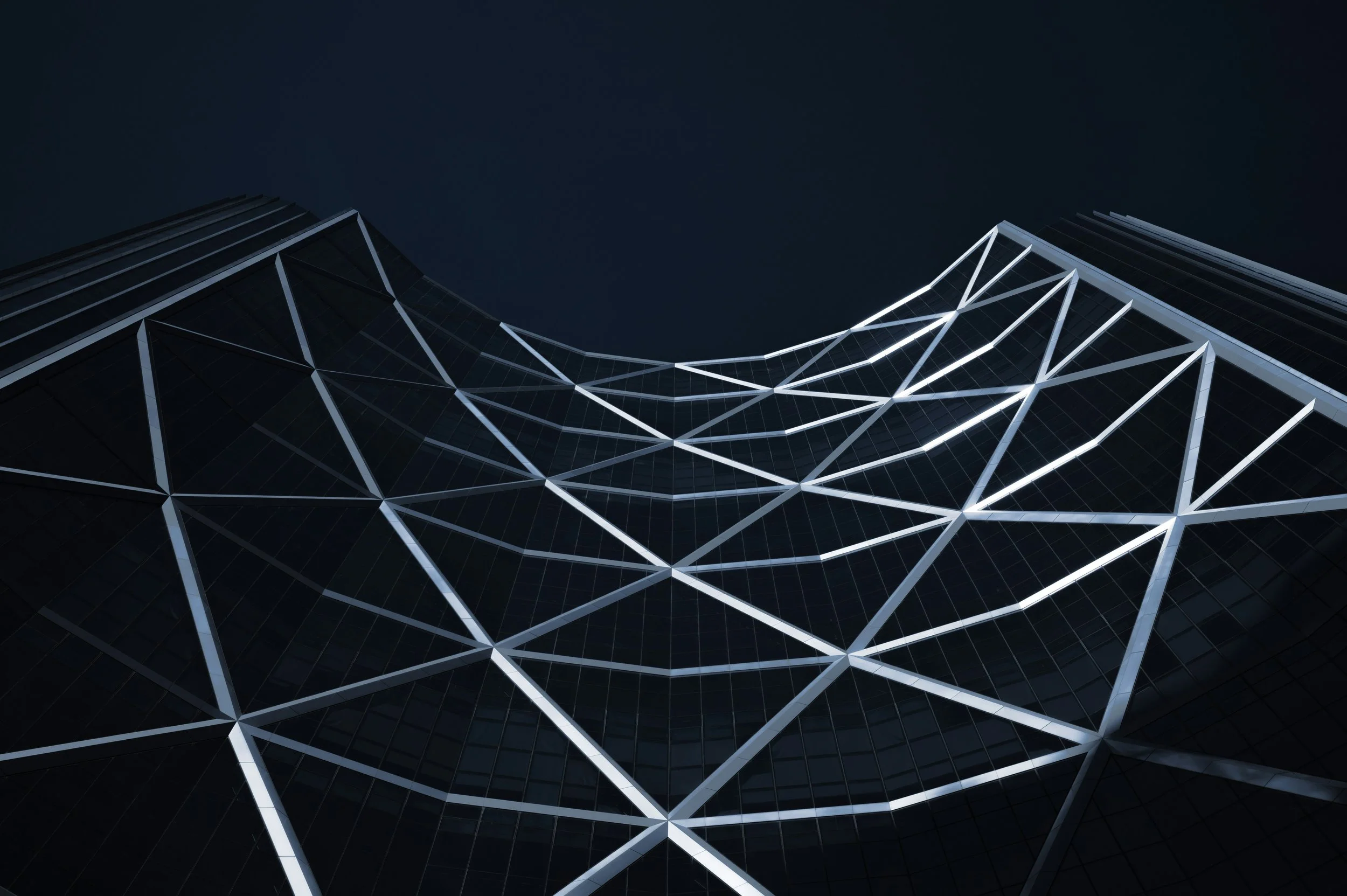 Low-angle view of a modern glass skyscraper at night, illuminated with white lights highlighting the geometric design.