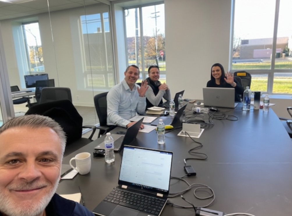 Four people sitting around a conference table in a bright office, waving and smiling at the camera. The table has laptops, water bottles, and notebooks.