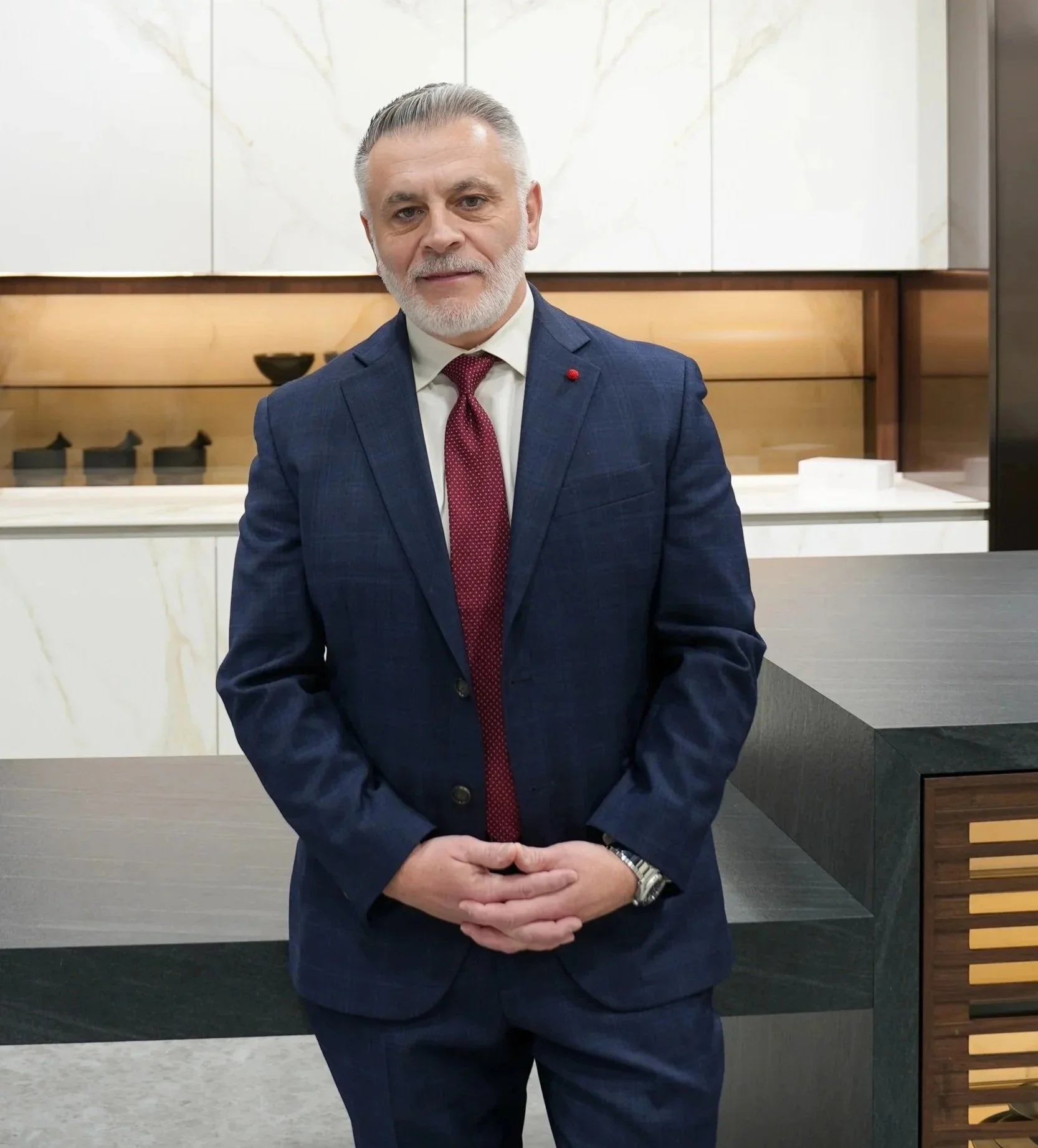 A man with gray hair and beard wearing a blue suit, white shirt, and red tie, standing in front of a modern, minimalist background with white marble and wood panels.