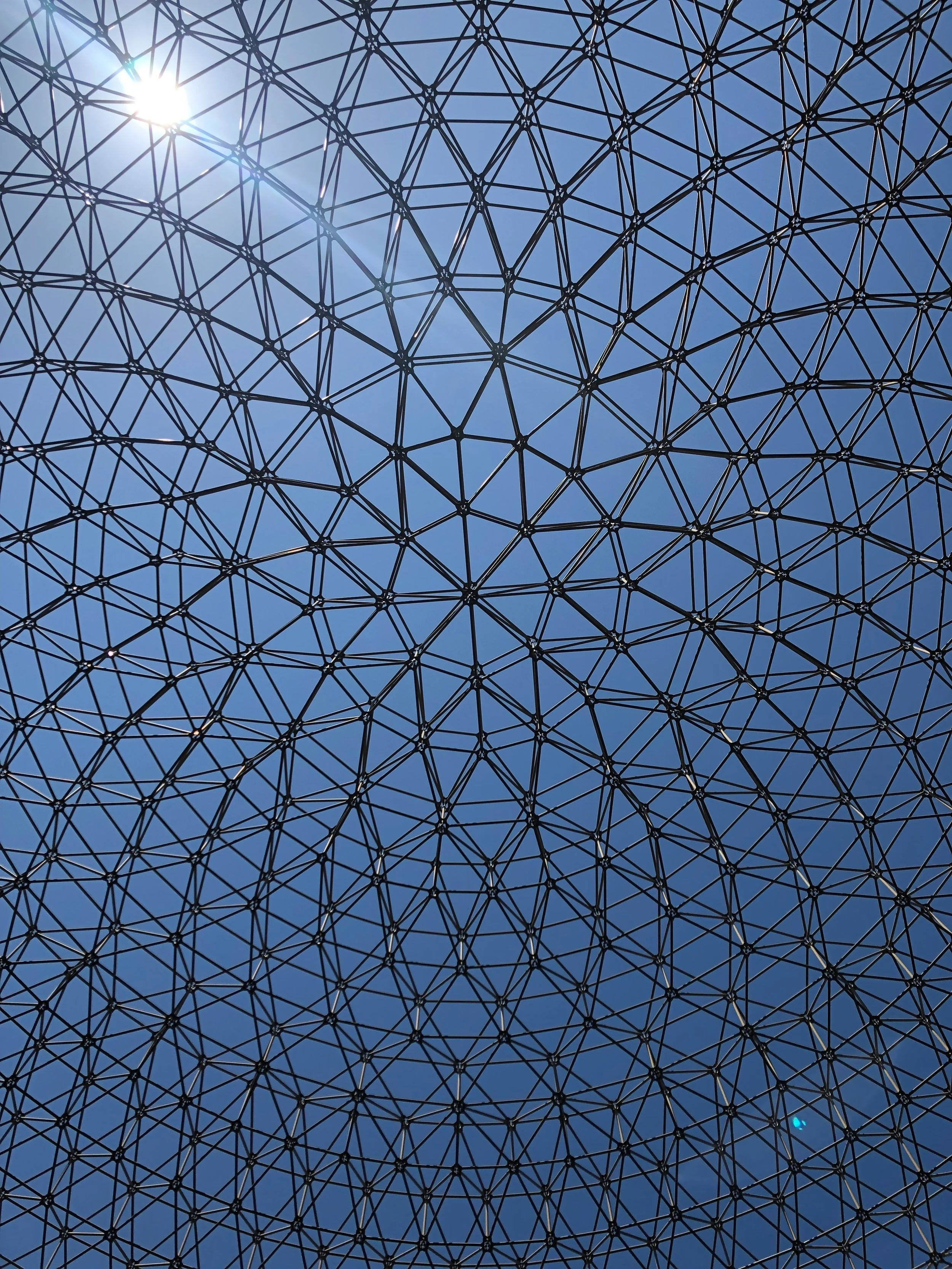 Looking up at a geodesic dome structure with a bright sun shining through the black metal framework against a clear blue sky.