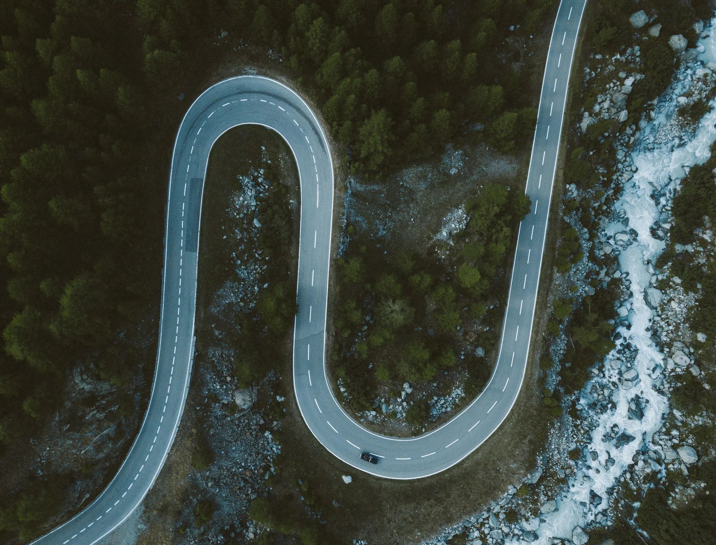 An aerial view of a winding mountain road with a car traveling along it, surrounded by trees, rocks, and a flowing river.