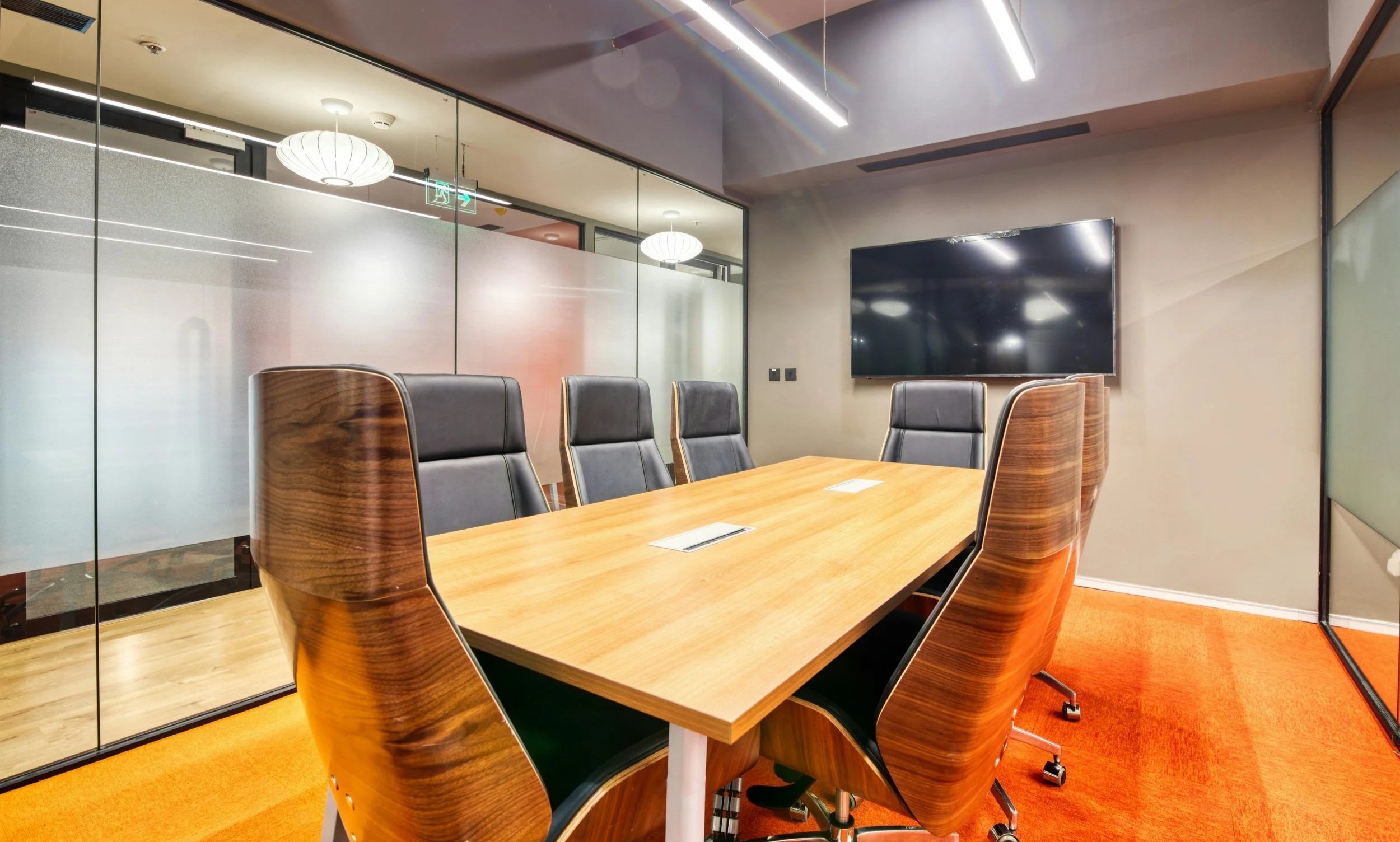 Modern conference room with wooden table, eight black leather chairs, a large wall-mounted TV, and frosted glass partitions.