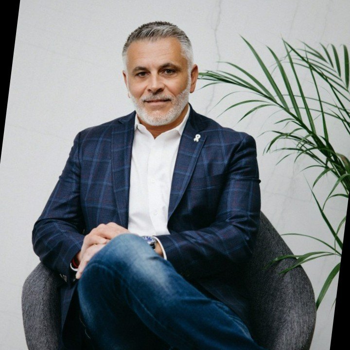 A middle-aged man with gray hair and beard sitting on a gray armchair, wearing a blue plaid blazer, white shirt, and dark jeans, with a small ribbon pin on his blazer, in front of a white wall with a green plant.
