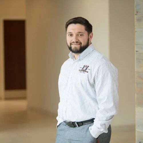 Portrait of a man with dark hair and a beard, wearing a striped button-up shirt with a logo and gray pants, smiling and standing indoors.