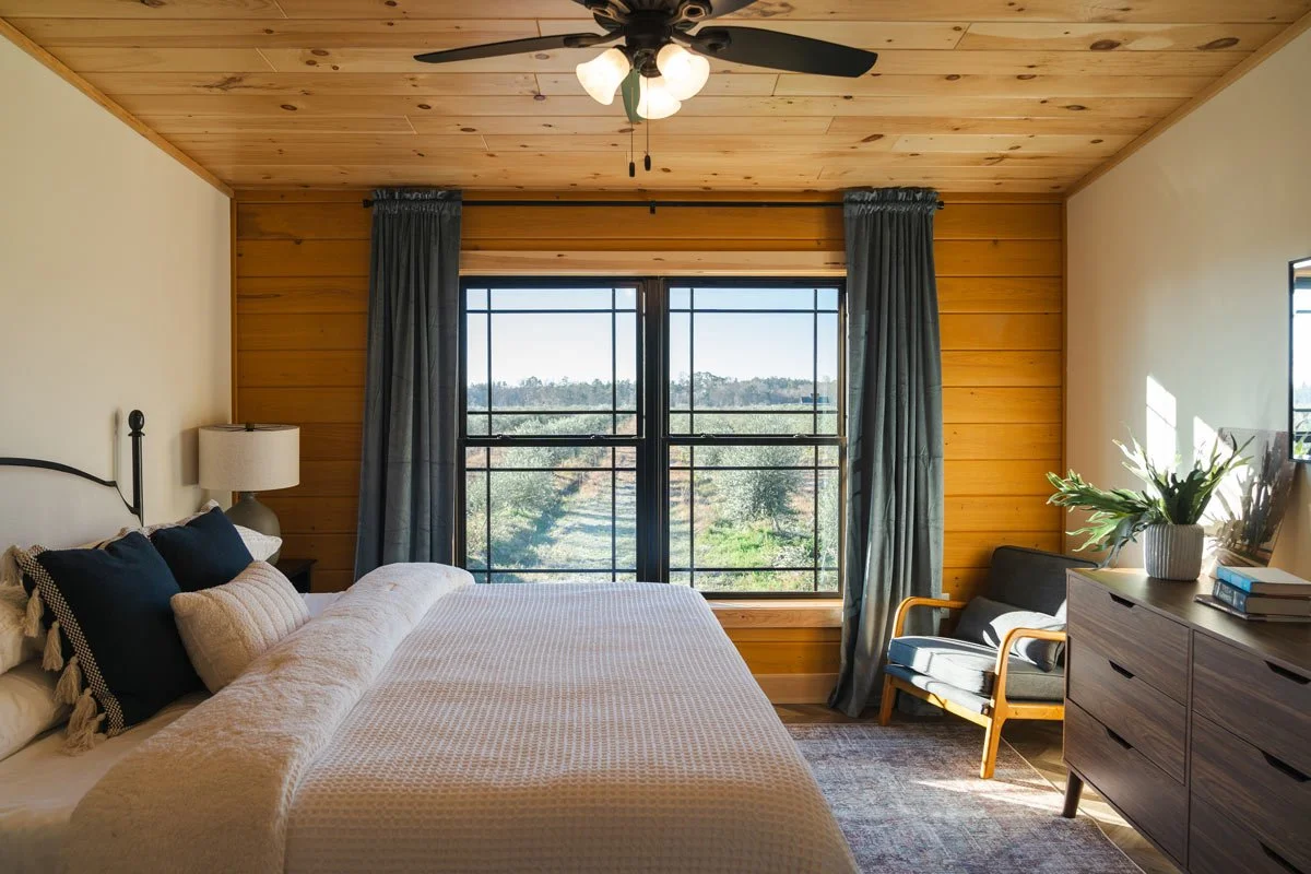 Cozy bedroom with wooden accents, large window with blue curtains, bed with pillows, side table with lamp, armchair, and dresser with plants and books.