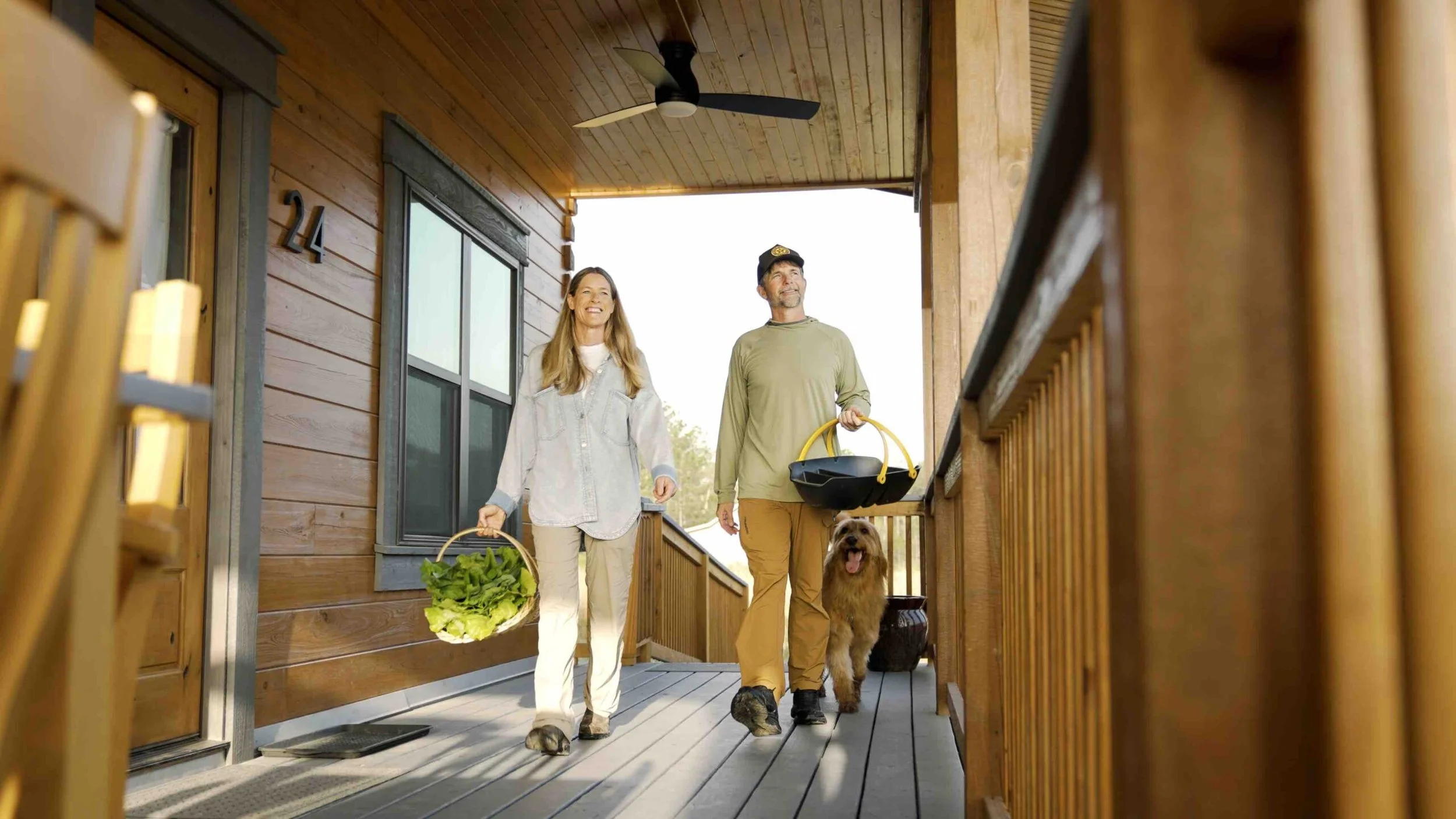 A man and woman walking on a wooden porch with a dog, holding baskets of greens and gardening tools, smiling and looking happy.