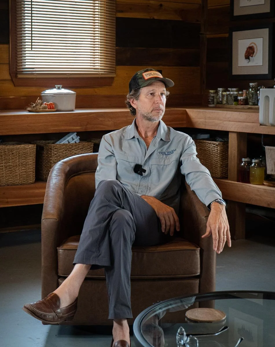 A man with gray hair and a beard sits in a brown armchair in a wooden-paneled room. He is wearing a gray work shirt, gray pants, a baseball cap, and brown slip-on shoes. He appears to be serious and focused, with one ankle resting on his knee. The background shows shelves with jars and baskets, a window with closed blinds, and framed pictures of roosters on the wall.