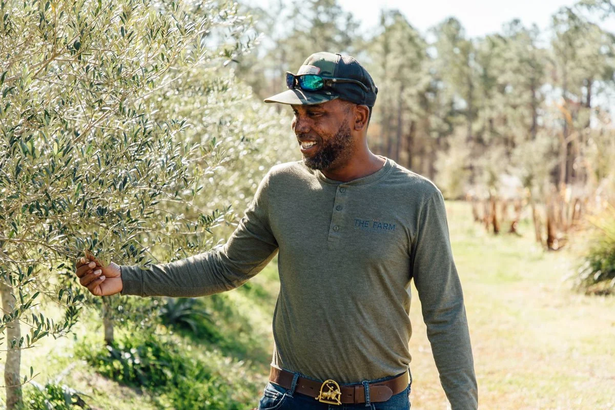 A man outdoors on a farm, standing near a tree and examining its leaves, wearing a long-sleeve shirt with 'THE FARM' on it, a cap, sunglasses on his head, and a belt with a gold buckle.