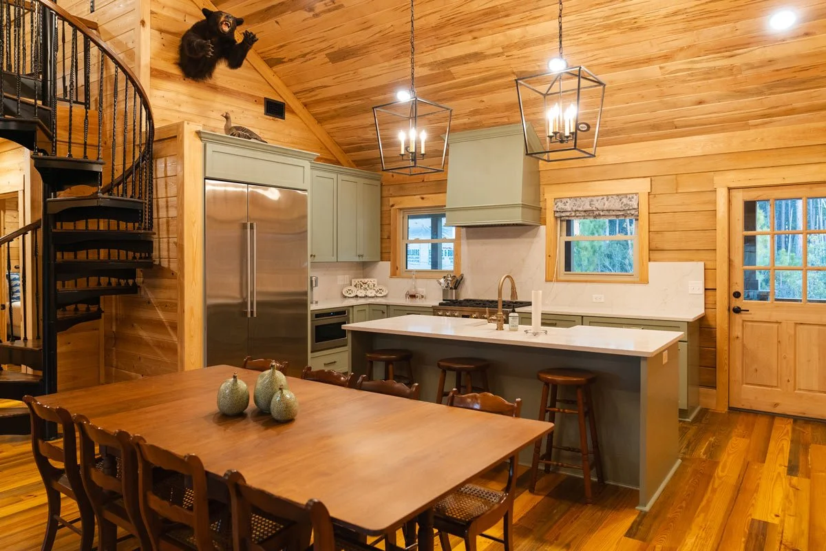 Open-concept kitchen and dining area with wood paneling, green cabinets, a kitchen island with stools, a wooden dining table with decorative vases, and a spiral staircase with a bear and a raccoon mounted on the wall.