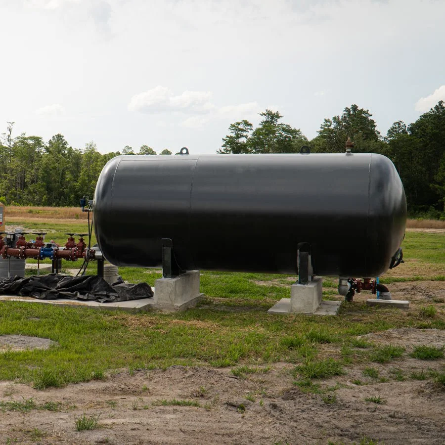 A large black propane tank installed outdoors on concrete blocks with piping and valves, set on a grassy field with trees and a cloudy sky in the background.