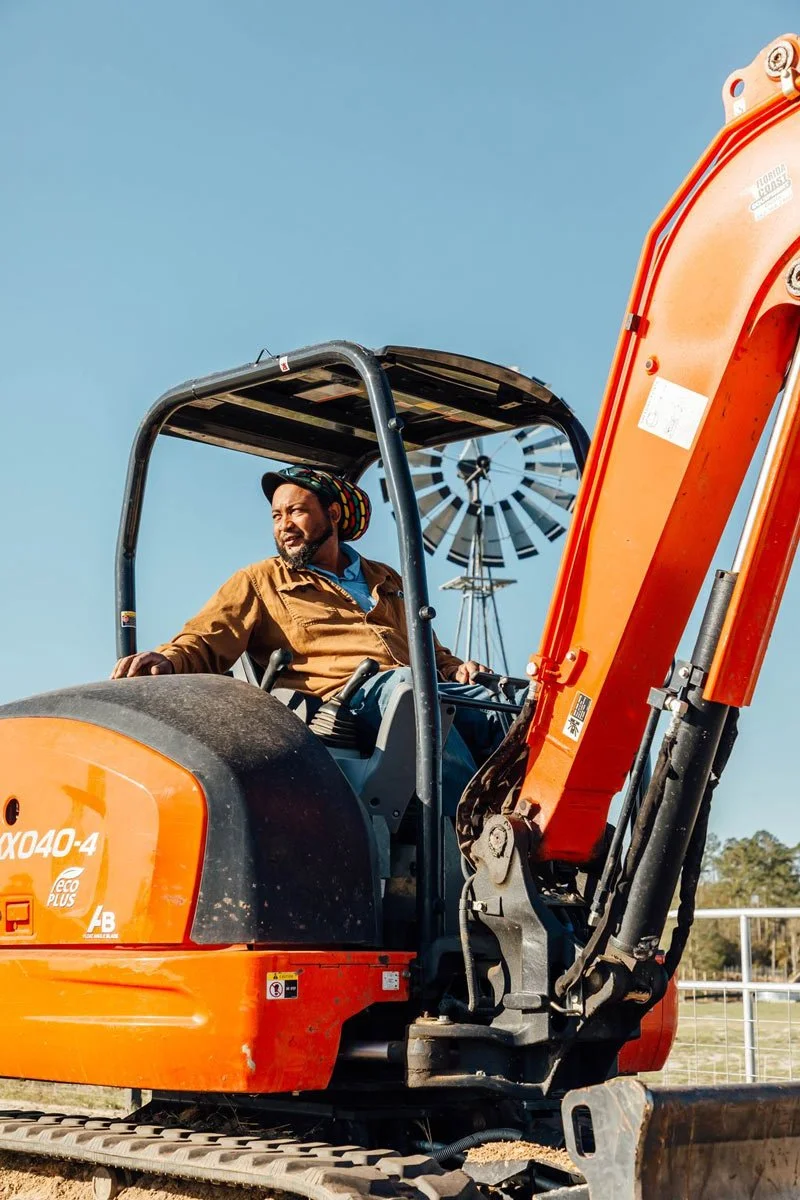 A man sitting in an orange excavator at a construction site, with a windmill and clear blue sky in the background.