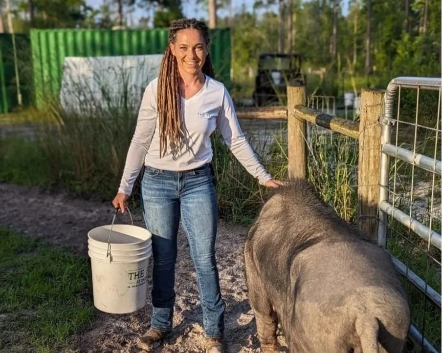 Woman with long braided hair holding a bucket and petting a pig on a farm or petting zoo. Green shipping container and trees in the background.