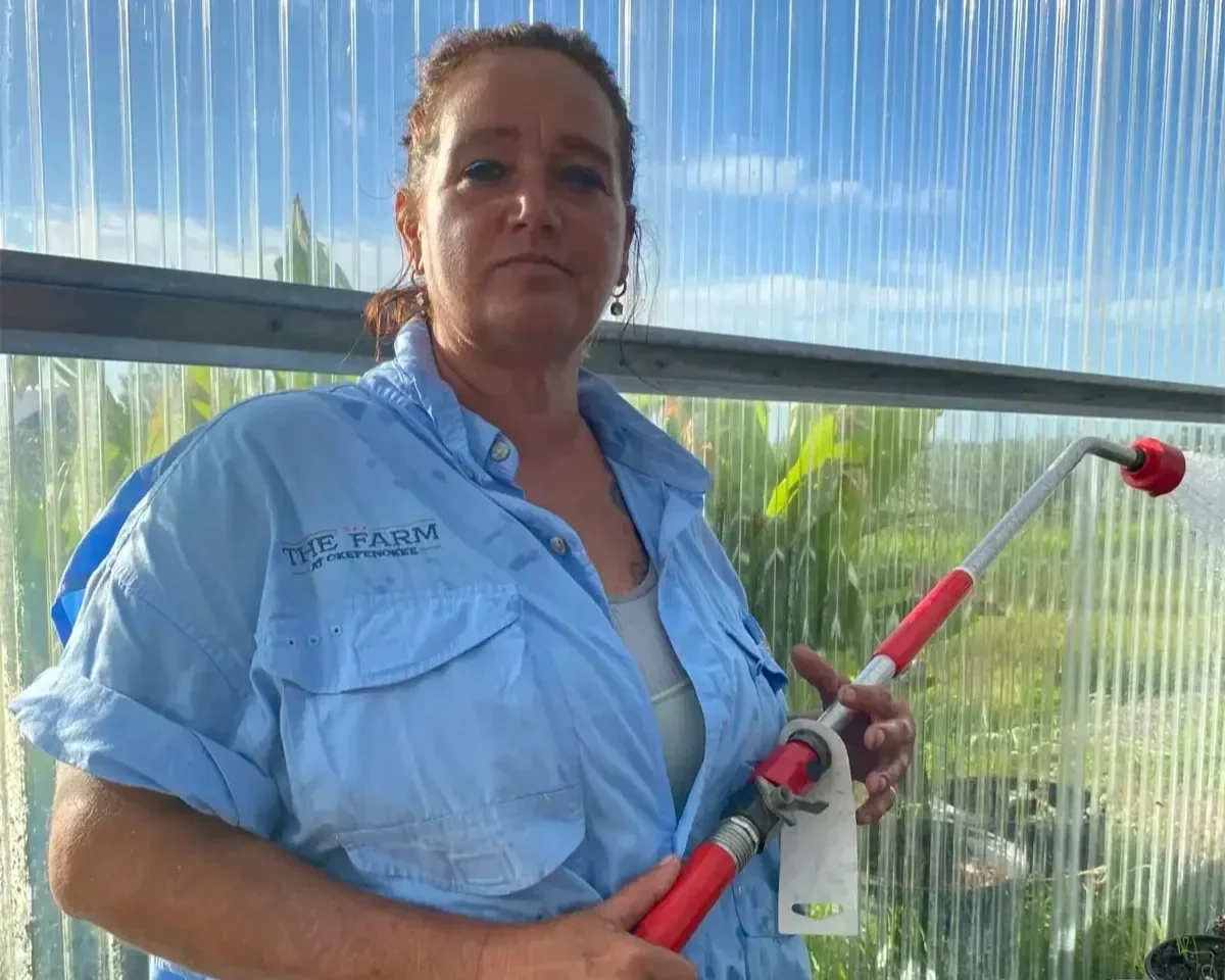 A woman wearing a light blue shirt holding a garden sprayer inside a greenhouse with green plants and a blue sky in the background.