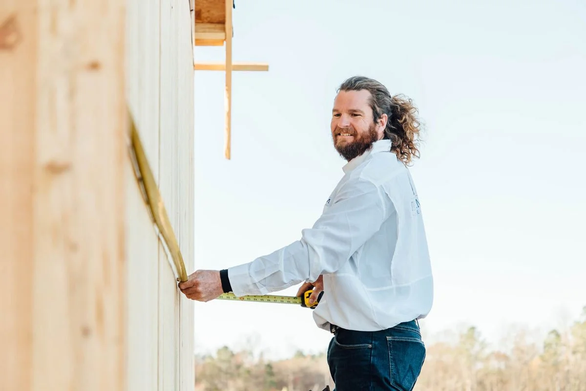 Man with curly hair and beard measuring a wooden wall with a tape measure at a construction site.