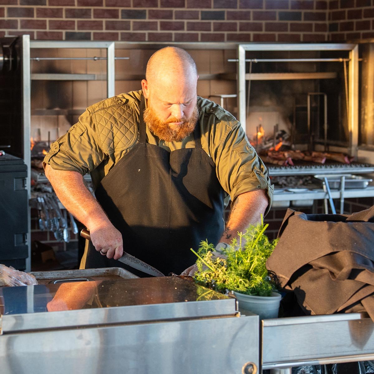 A man with a beard in a green shirt and black apron is chopping herbs on a stainless steel counter in a professional kitchen with a brick wall and open flame grills in the background.