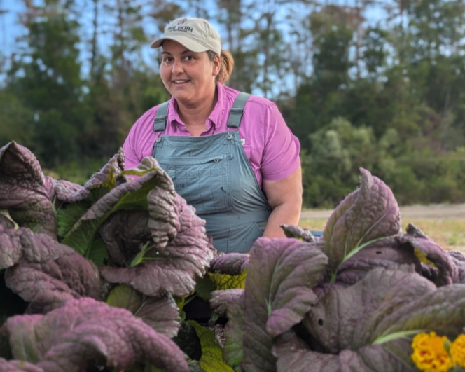 A woman in purple shirt and overalls smiling in a garden, surrounded by purple and green leafy plants with a background of trees.