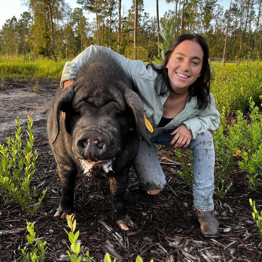 A woman smiling and crouching next to a large black pig in a muddy, green outdoor area with trees and plants.