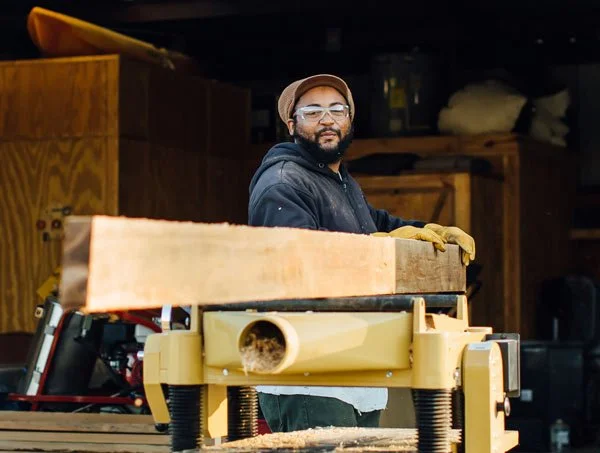 A man in safety glasses, a hat, and gloves working in a woodworking shop.
