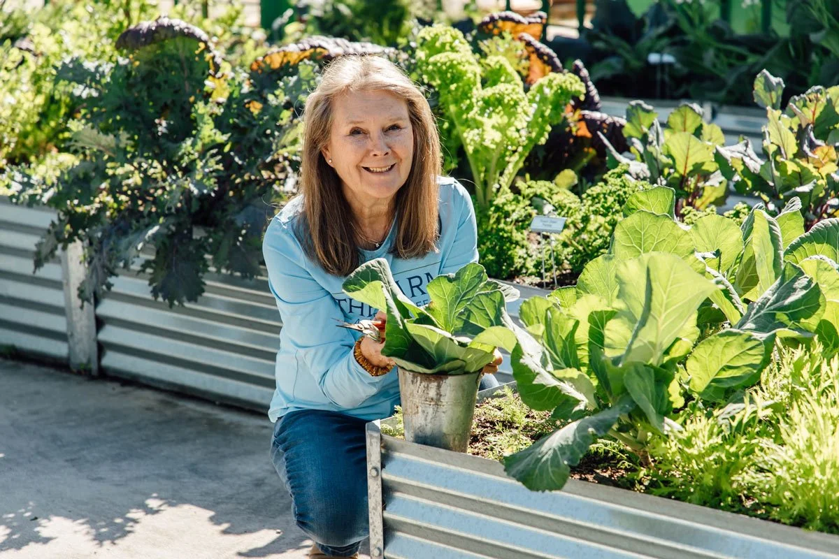 A smiling woman in a light blue shirt kneeling next to a potted lettuce plant in an outdoor garden or farm with various leafy greens.