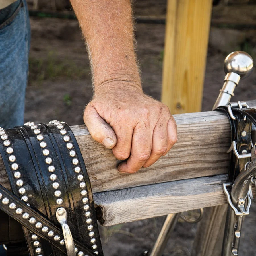 Close-up of a person's weathered hand gripping a wooden beam, with motorcycle handlebar and leather straps with metal studs in the background.