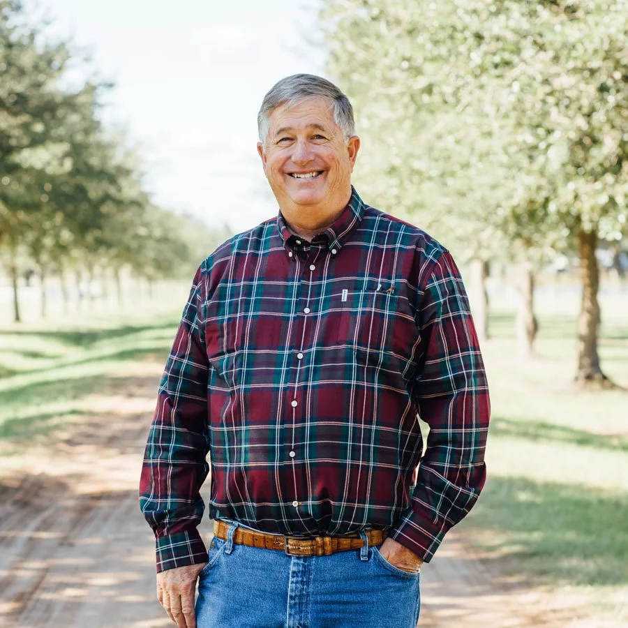 A smiling older man with gray hair wearing a plaid shirt and jeans stands outdoors on a sunny day, with trees and a dirt path in the background.