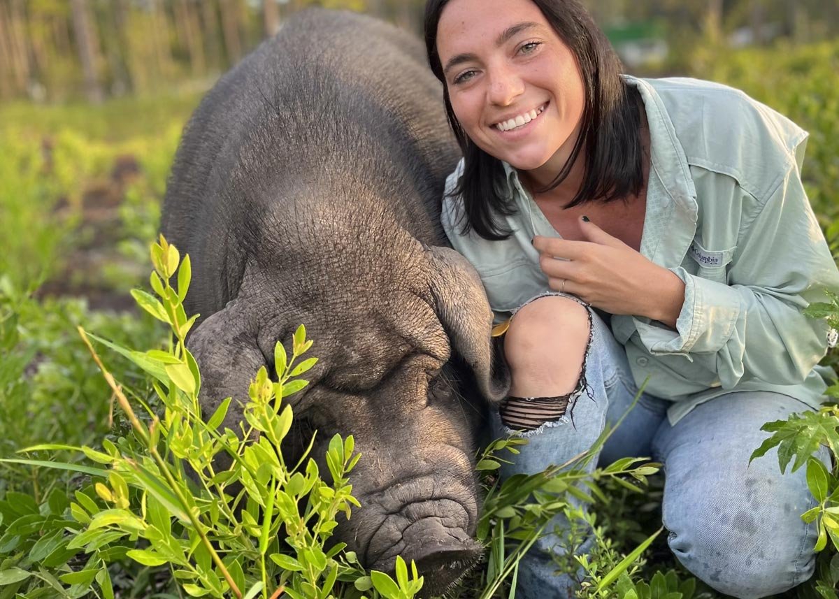 A woman kneeling next to a sleeping baby elephant in a grassy field, smiling at the camera.
