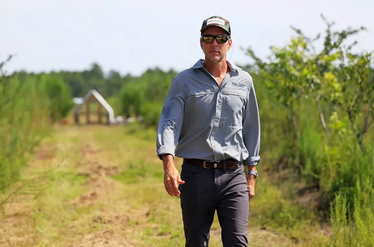 A man wearing sunglasses, a baseball cap, a gray long-sleeve shirt, and dark pants walking in a field with green plants on either side and a small house in the background.