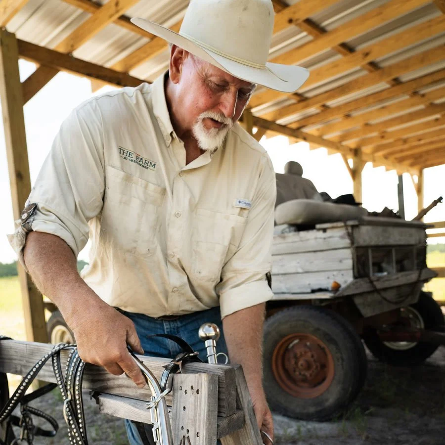 An elderly man wearing a hat and a shirt with 'The Farm' written on it is working on a woodworking project under a wooden shelter. There is an old truck in the background.
