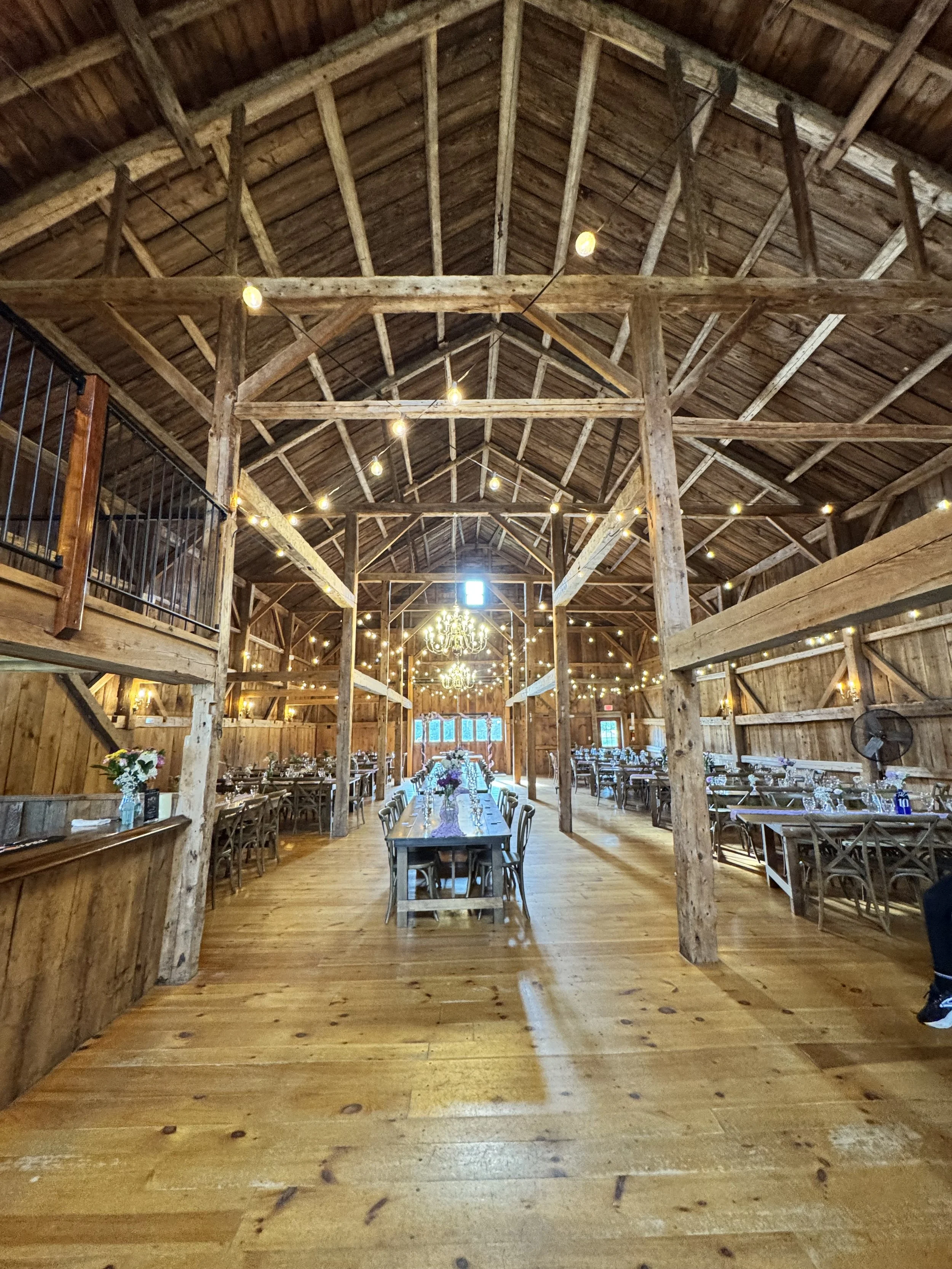 Interior of a rustic barn set up for an event with long tables, chairs, and string lights hanging from the wooden beams.