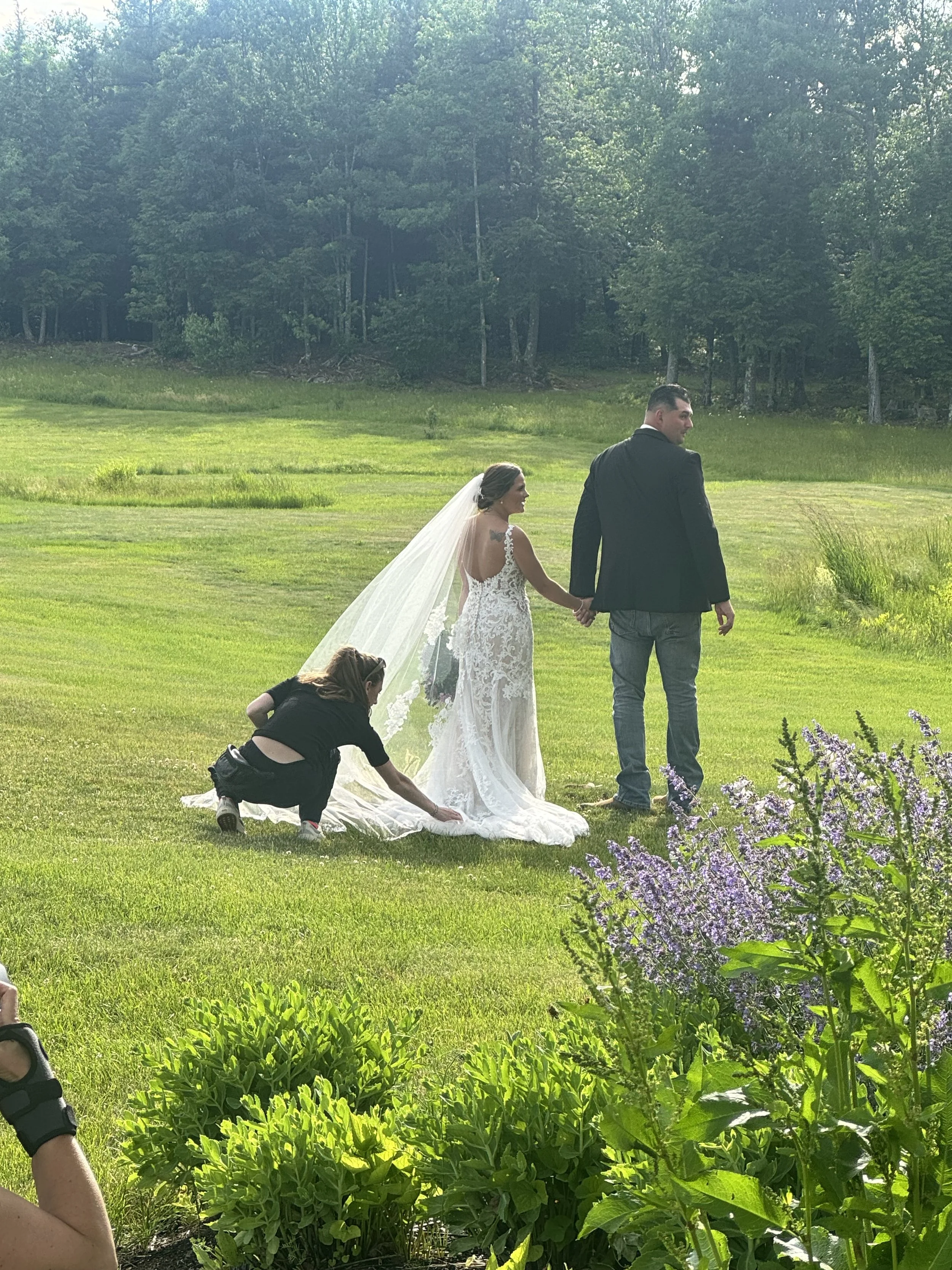 A bride and groom holding hands while walking on a grassy field with a woman crouching nearby. The bride is wearing a white lace wedding dress with a long veil, and the groom is in a dark suit with jeans. The background features trees and a bright, sunny sky.