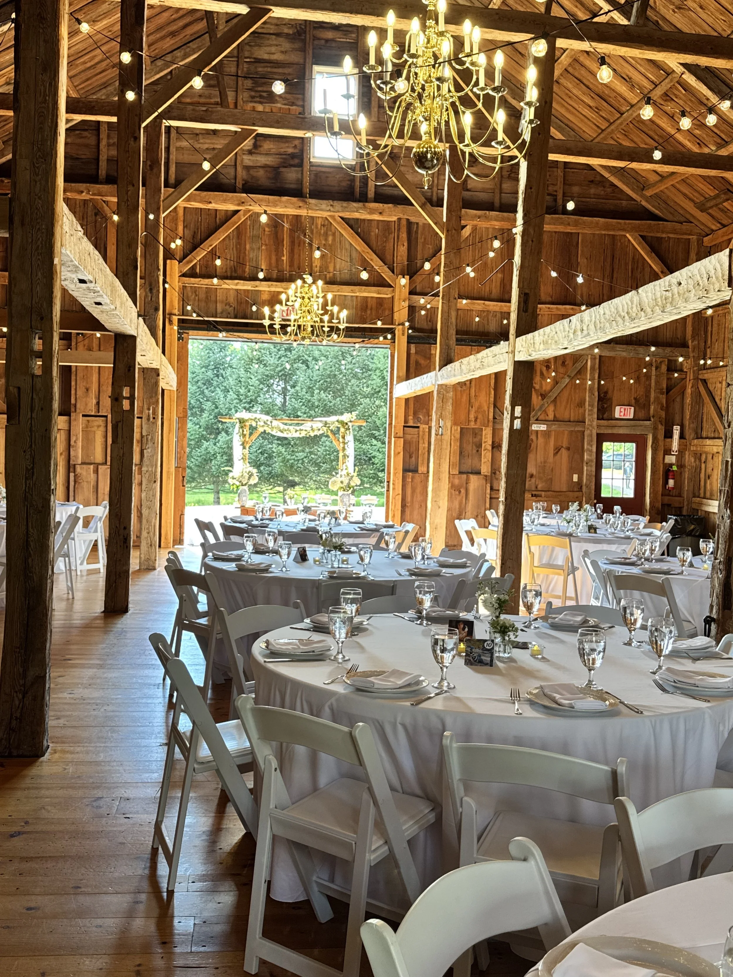 Indoor reception hall with wooden beams and walls, decorated with string lights and two chandeliers hanging from the ceiling, with round tables set with white tablecloths, plates, glasses, and silverware.