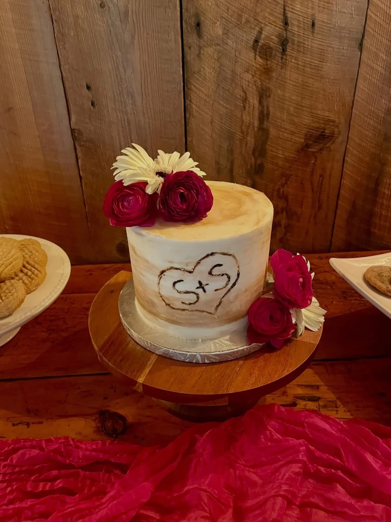 A wedding cake with a white frosted surface, decorated with pink and white flowers on top, and a heart with initials 'S + S' drawn in chocolate on the front. The cake is on a wooden table with a red fabric underneath and other plates of cookies nearby. The background is wooden paneling.