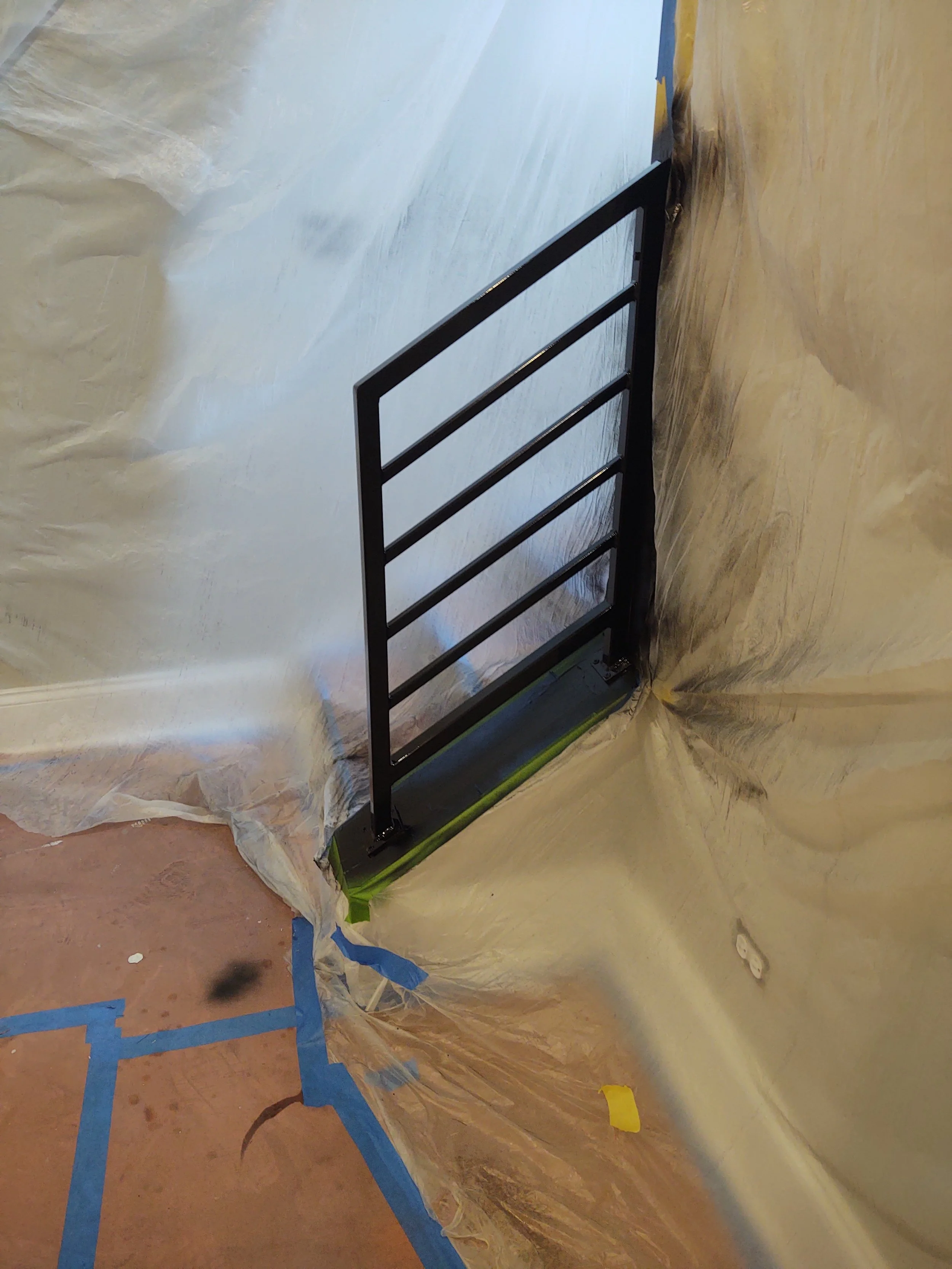 Black metal railing installed on a wall, surrounded by protective plastic and painter's tape during construction or renovation.