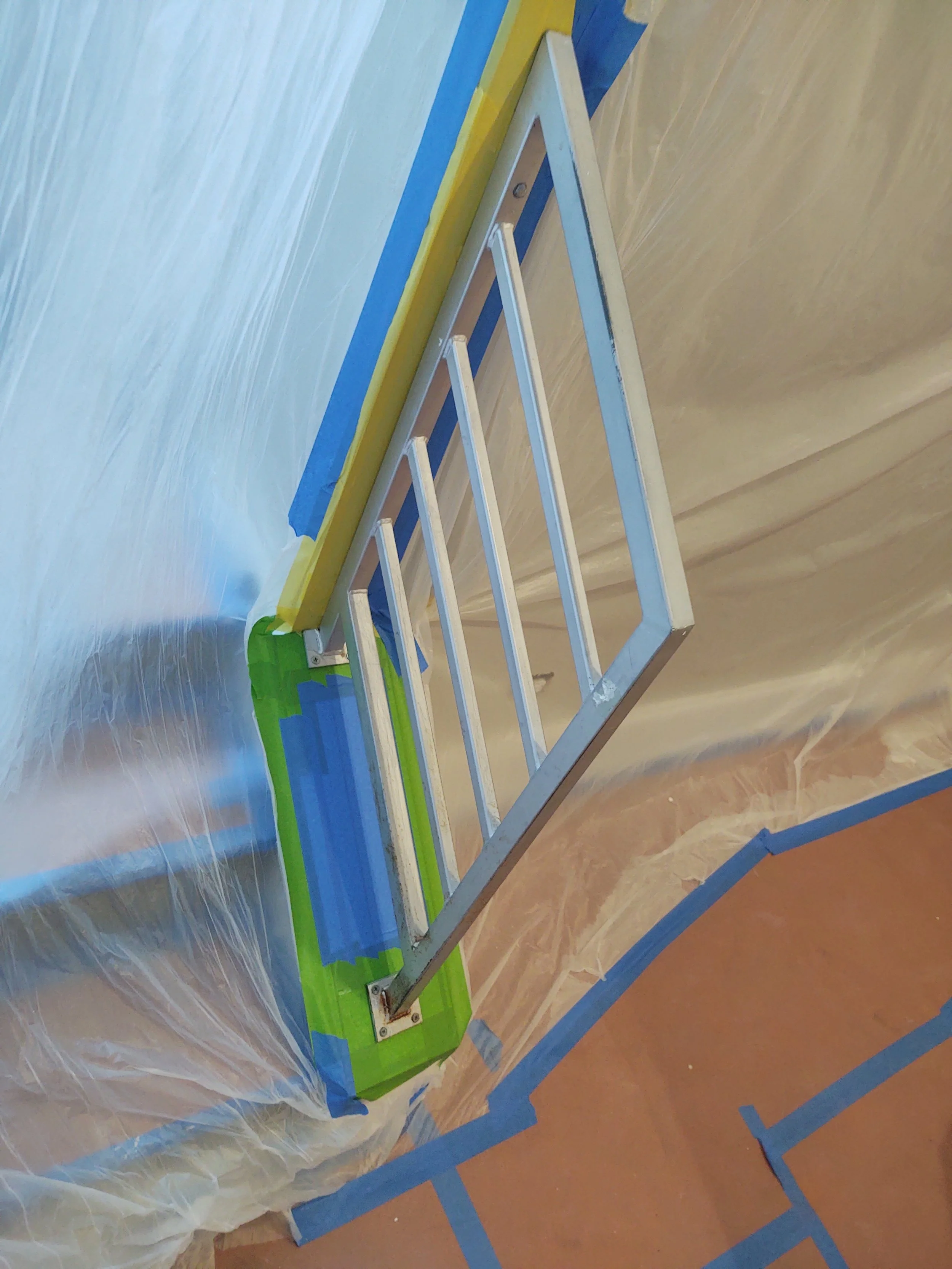 Silver metal ladder leaning against a wall covered with plastic sheeting during painting prep, with painter's tape on the floor and wall edges.