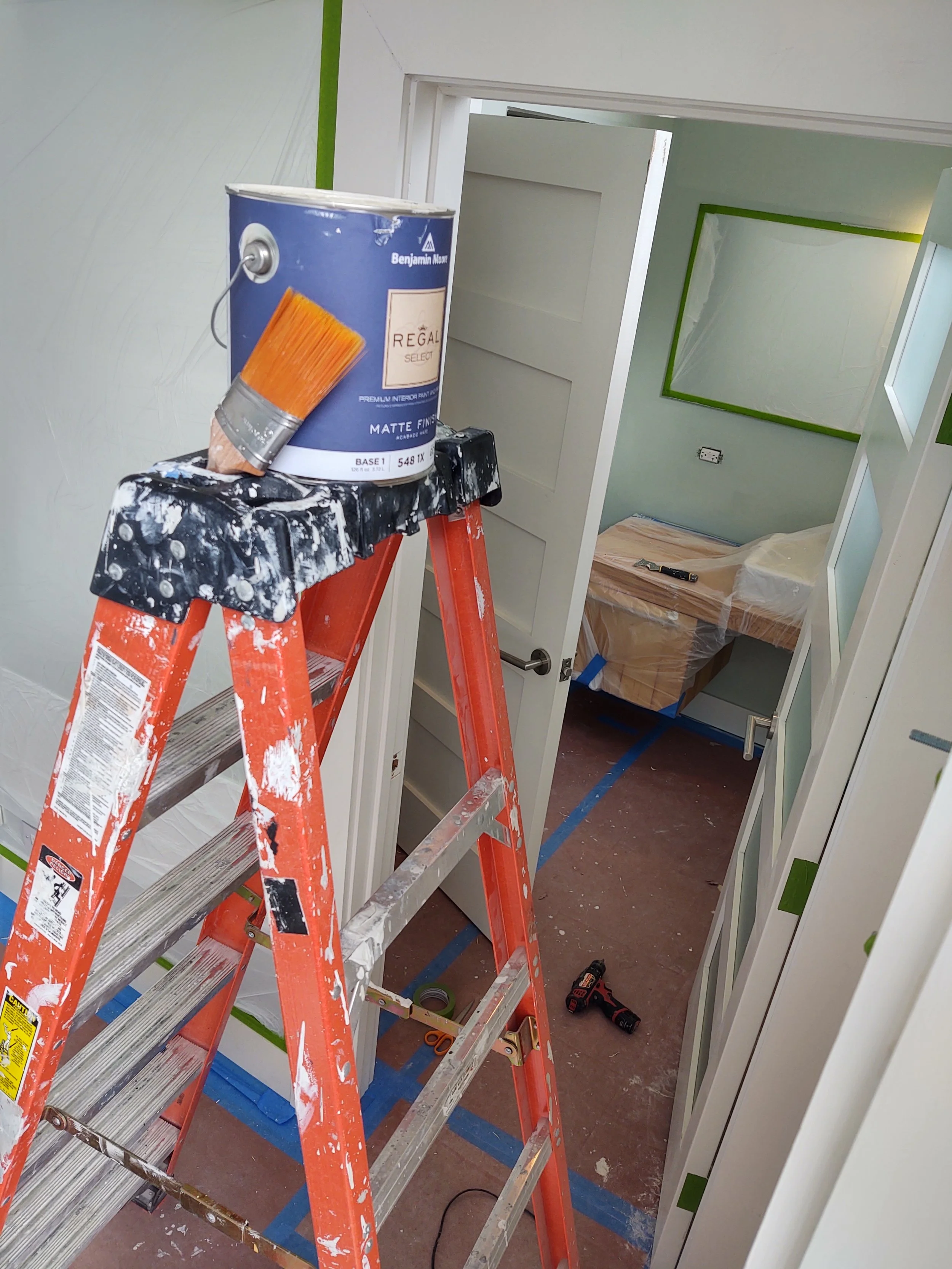 A ladder with paint splatters, a can of Benjamin Moore paint with a brush attached, a drill on the floor, and a room under renovation with a bed covered in plastic, blue painter's tape on the floor, and partially painted walls.