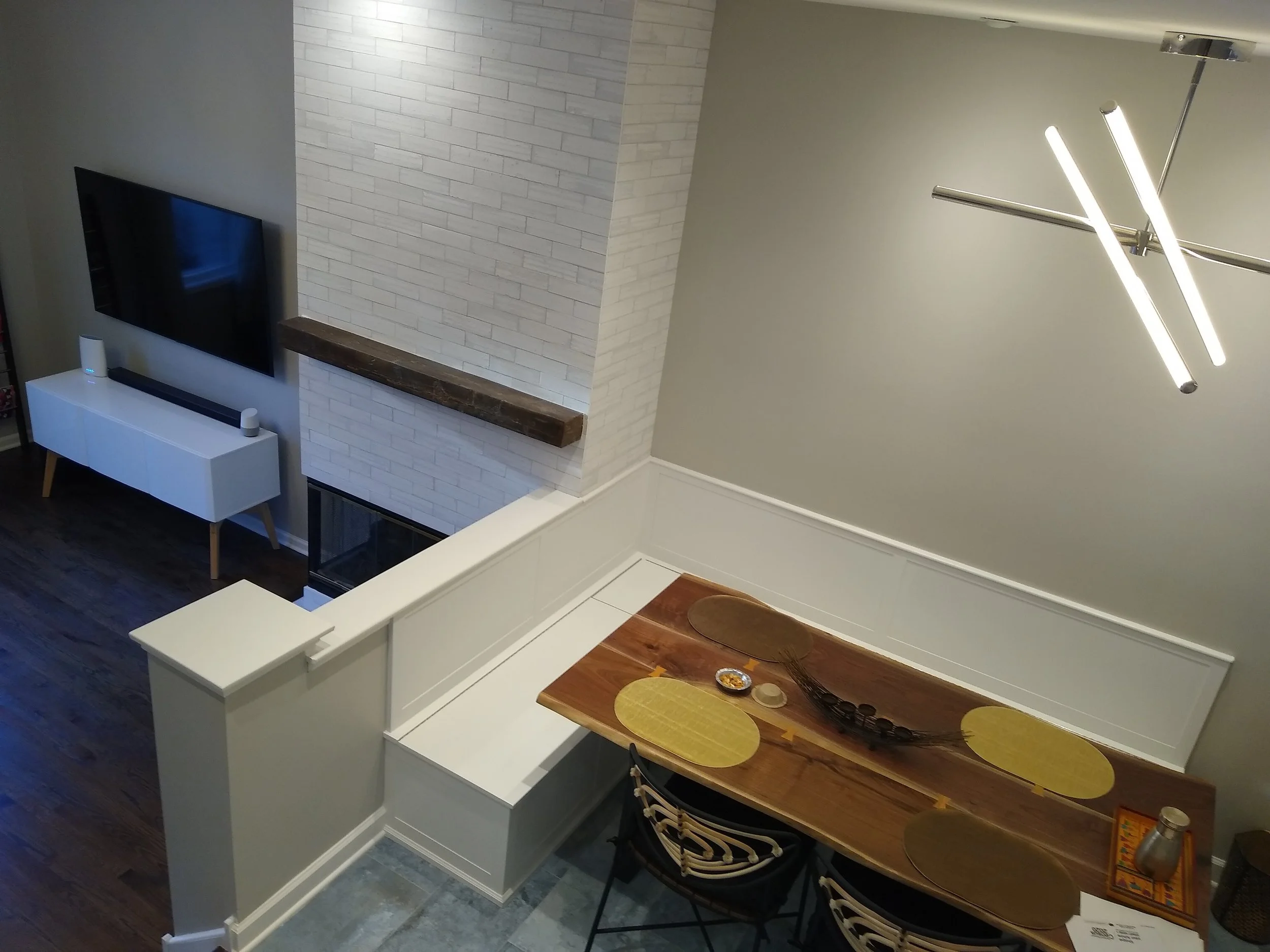 Interior view of a living and dining area with a wall-mounted TV, white entertainment console, fireplace, modern chandelier, wooden dining table with yellow placemats, and decorative bowl, with hardwood flooring and neutral wall colors.