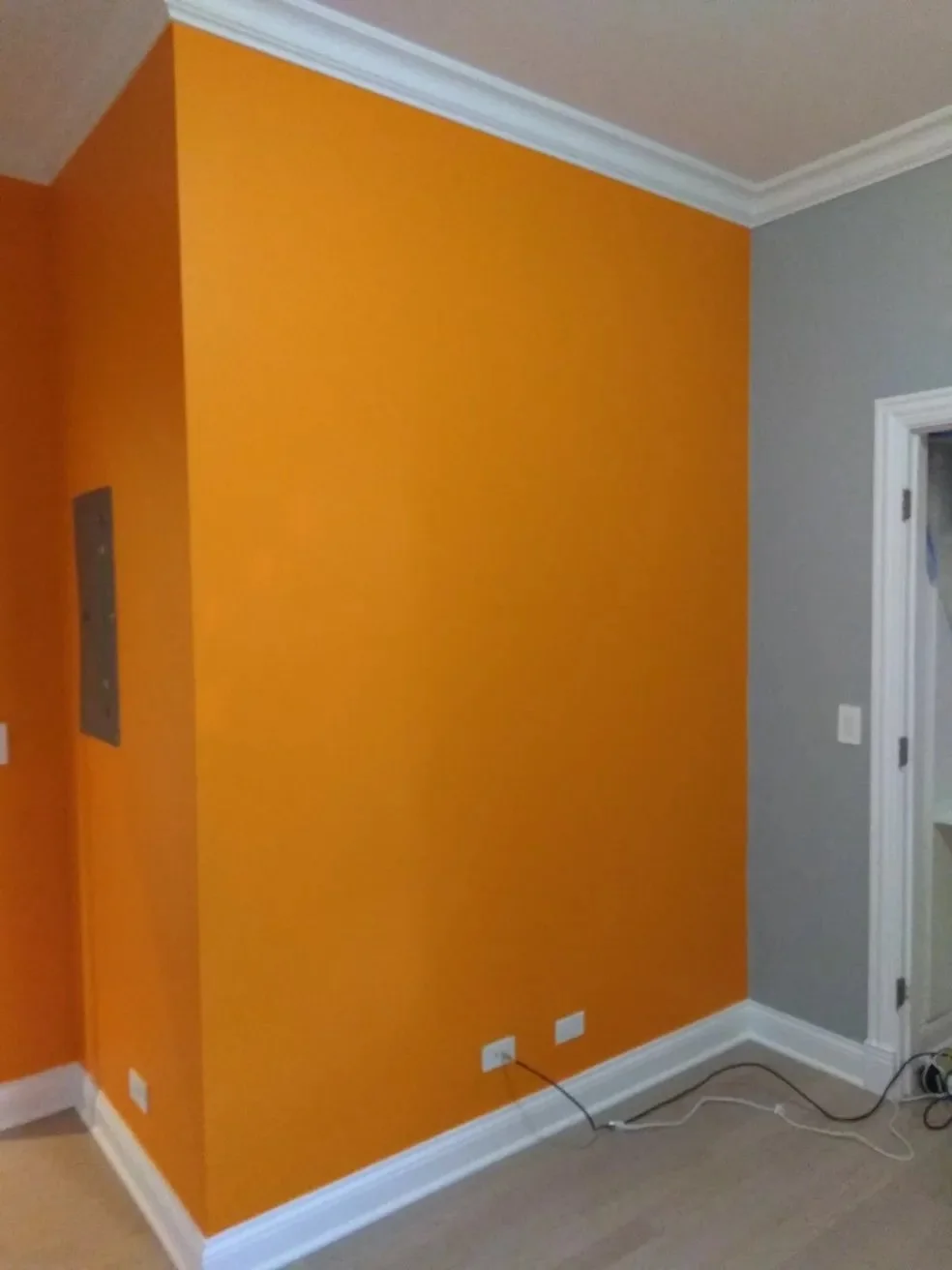 Interior corner of a room with a bright orange accent wall, white crown molding, gray wall, and white baseboard, with electrical outlets and cables on the floor.