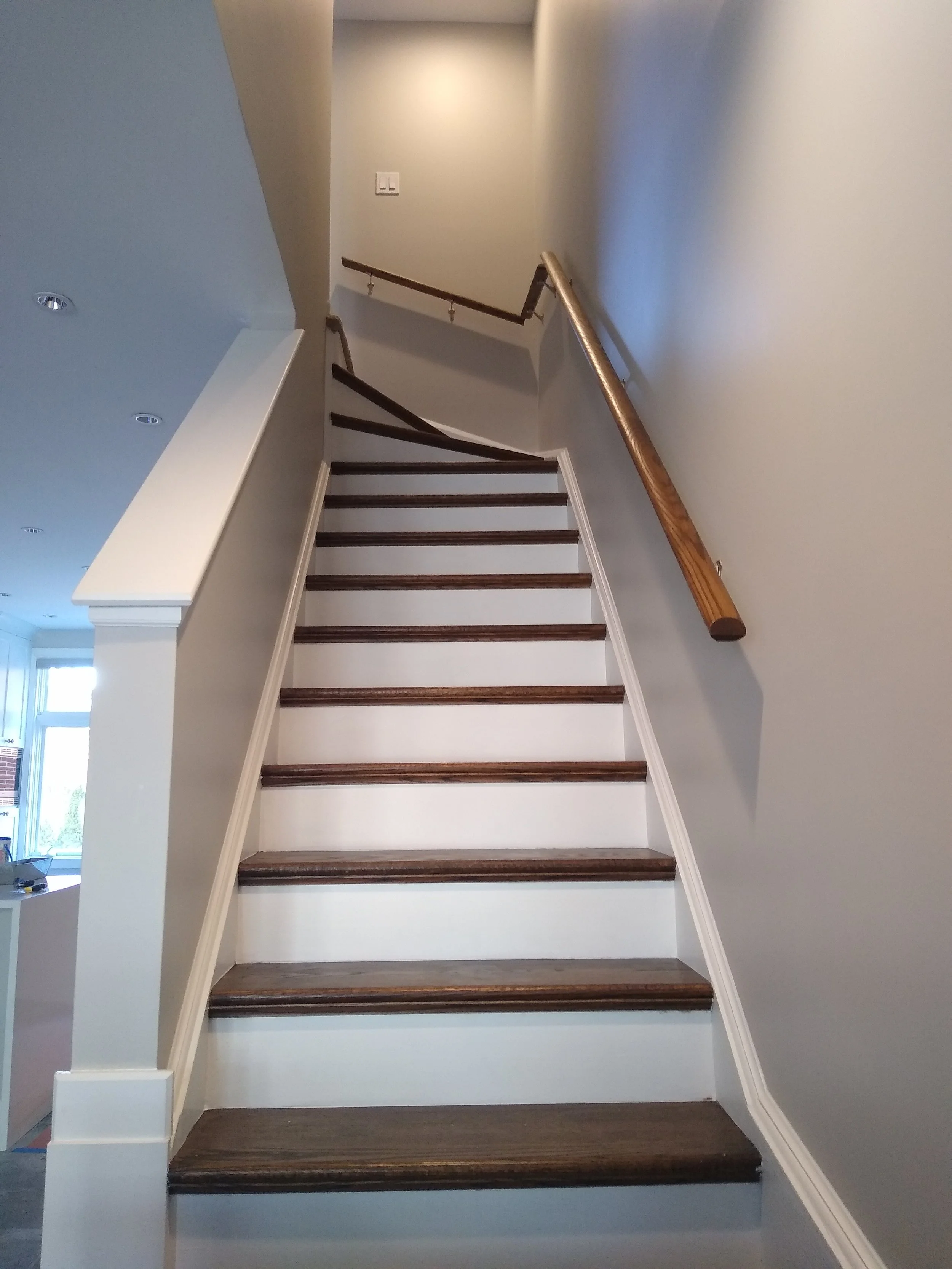 Indoor staircase with dark wooden steps and a wooden handrail on the right side, leading up to a landing with a beige wall and an electrical outlet, in a house.